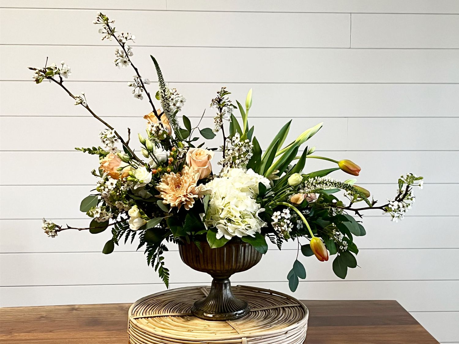 Classic bronze urn arrangement with hydrangeas, lilies and mixed greenery on wooden table.