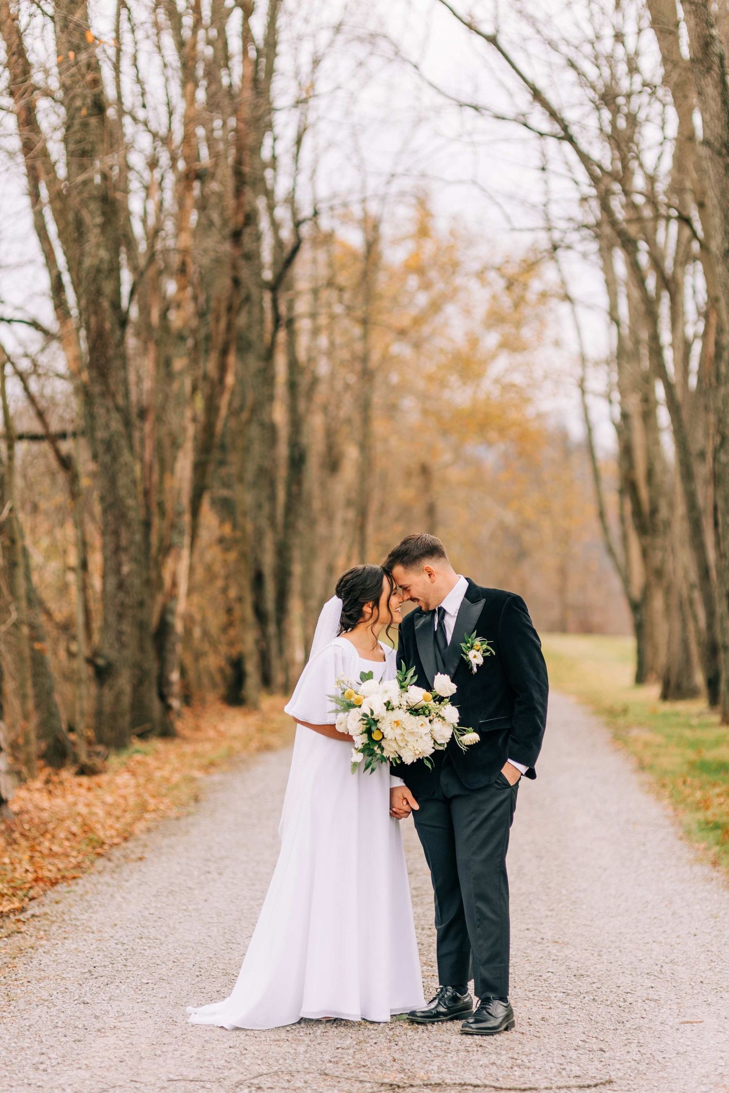Newlyweds holding bouquet pose together on autumn path lined with bare trees.