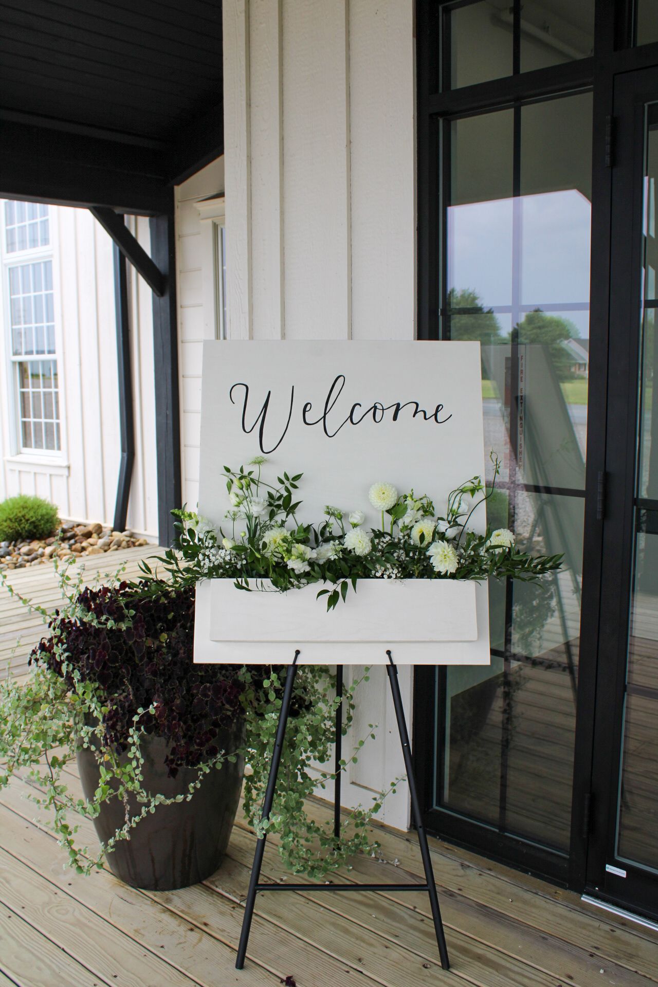 A rustic white welcome sign decorated with greenery and white flowers on an easel outside a venue.