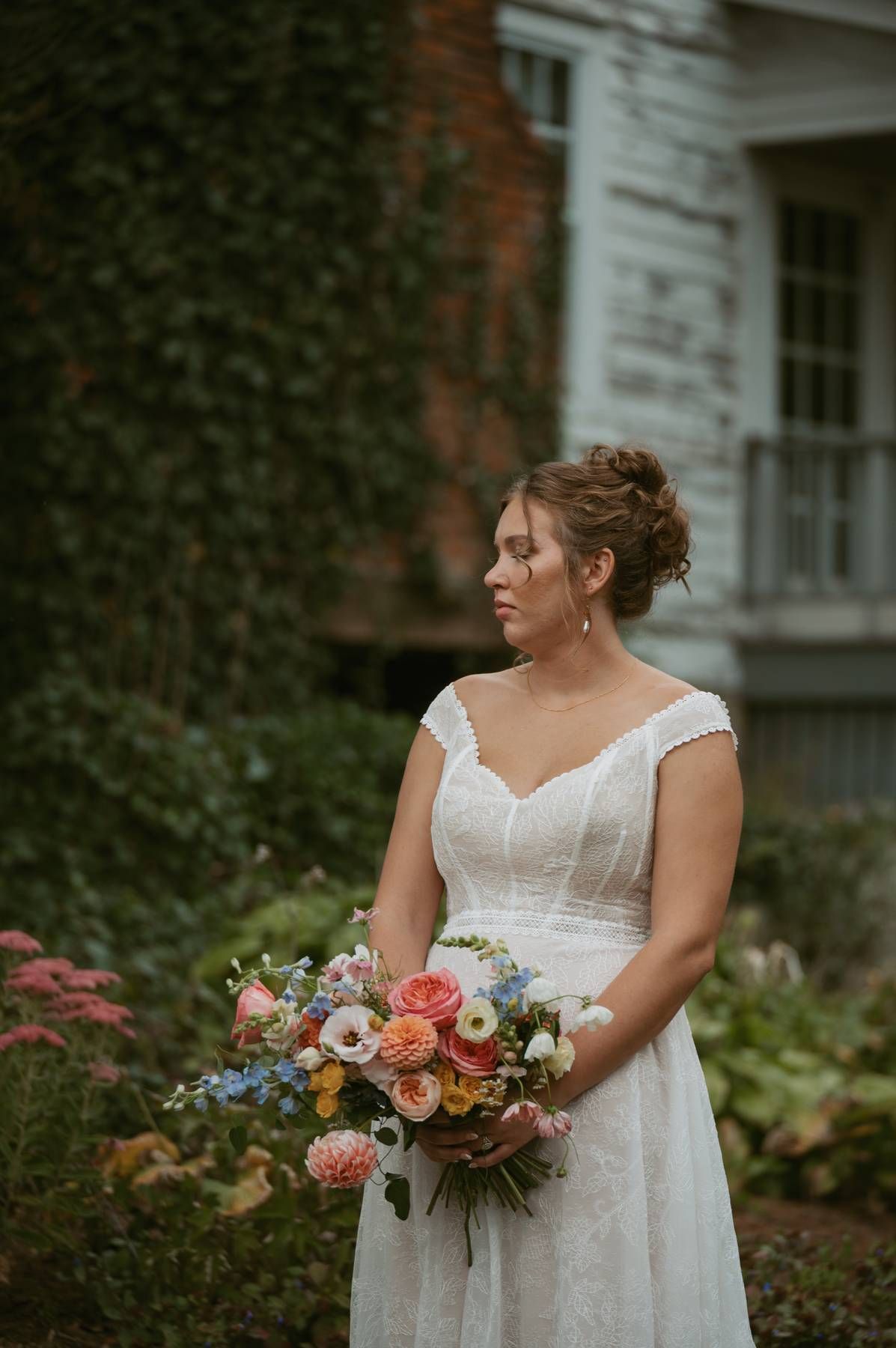 A bride in a white lace dress holds a vibrant bouquet of coral and blue flowers in a garden setting.