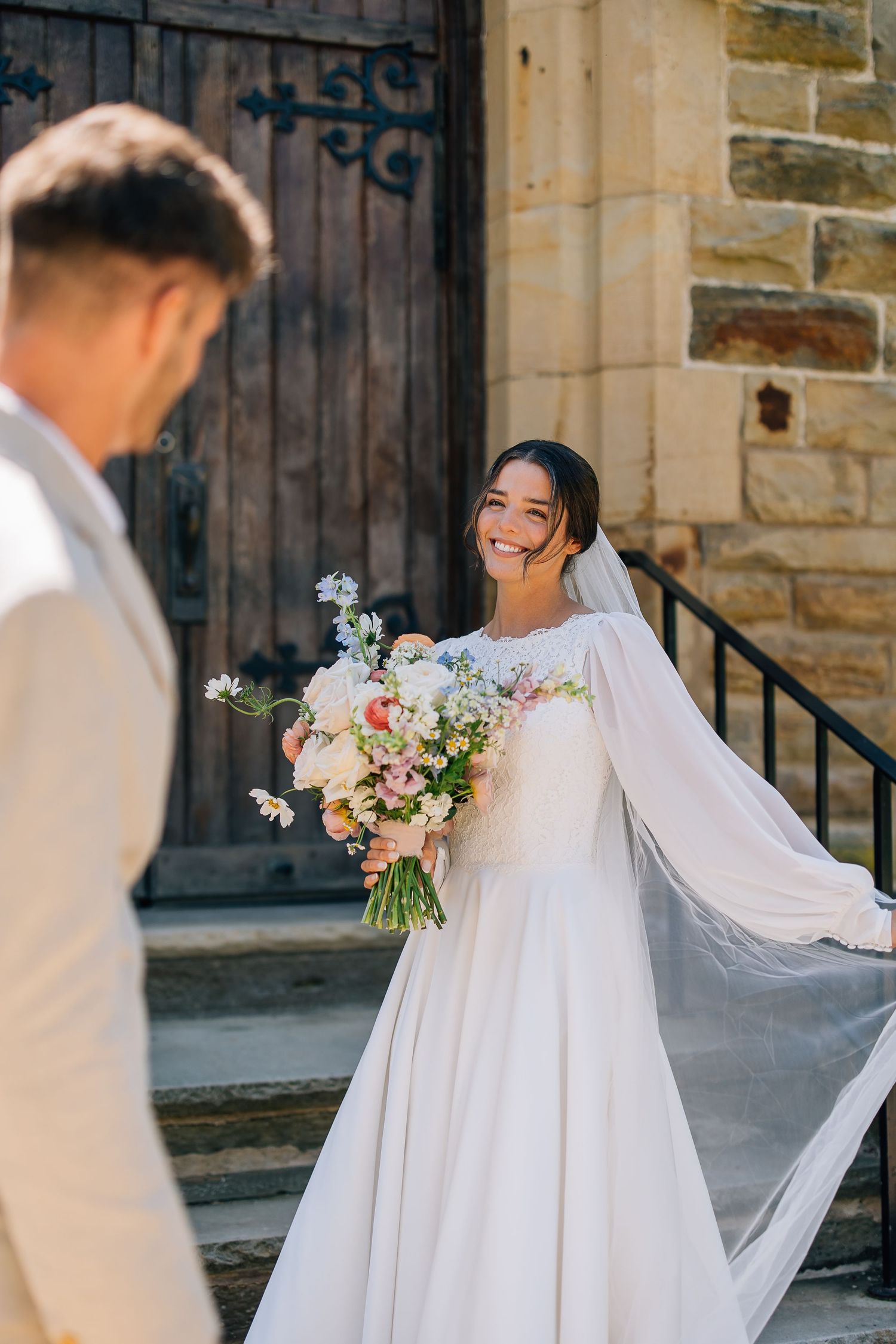 A bride in a flowing white wedding dress with a long veil holds a pastel bouquet outside a stone church.