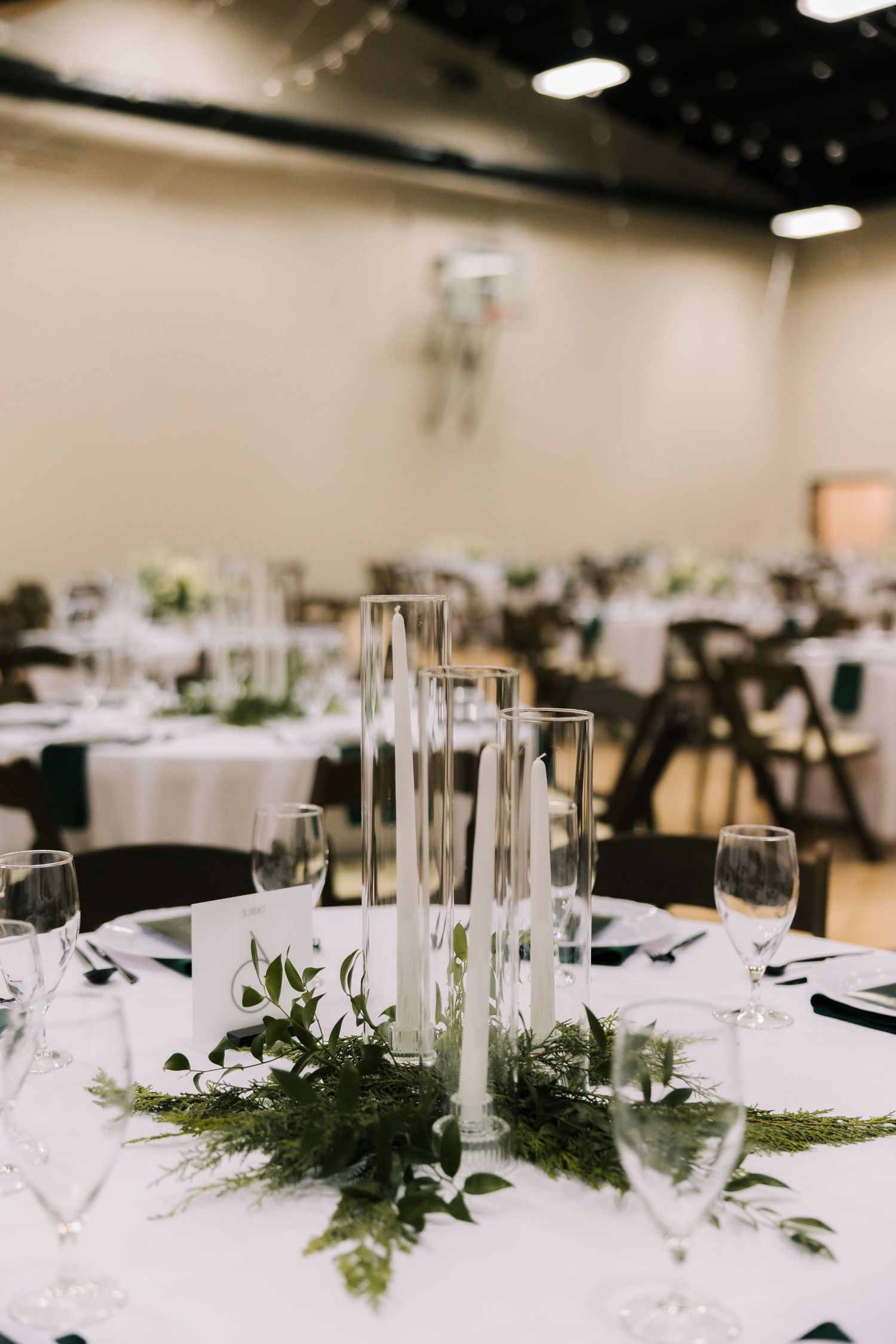 White table settings with evergreen foliage centerpieces and glass candle holders create an elegant reception atmosphere.