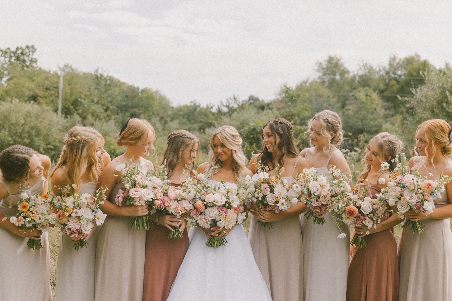 Wedding party in neutral tones stands together outdoors holding pastel floral bouquets.
