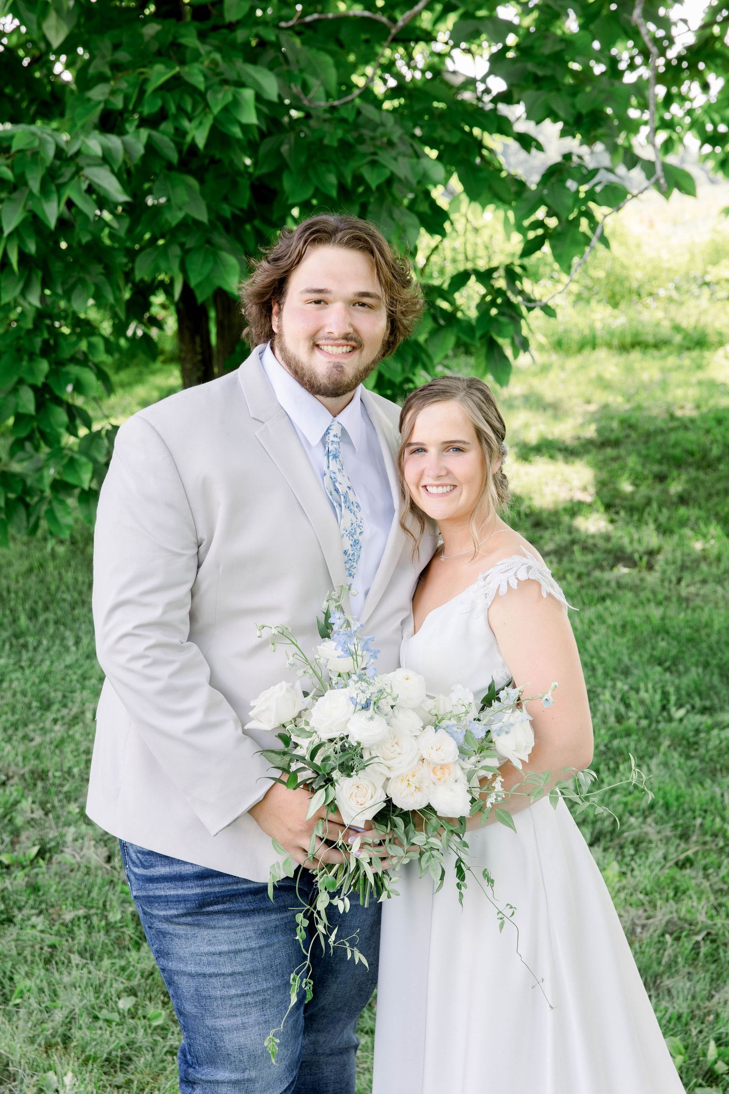 Casual country wedding portrait with couple holding white and green floral bouquet under tree.