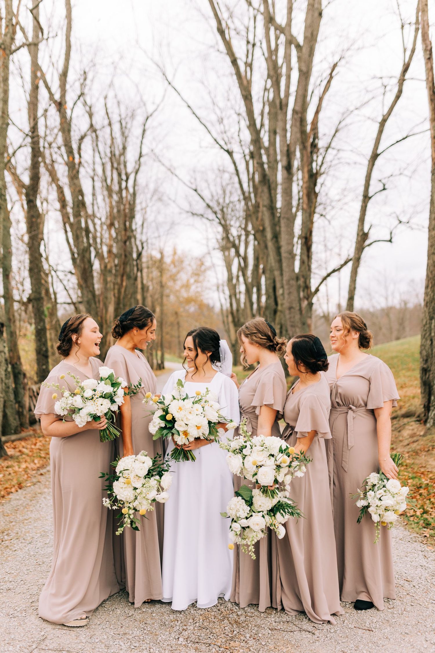 Wedding party in neutral mauve dresses poses on autumn path holding white and cream bouquets.