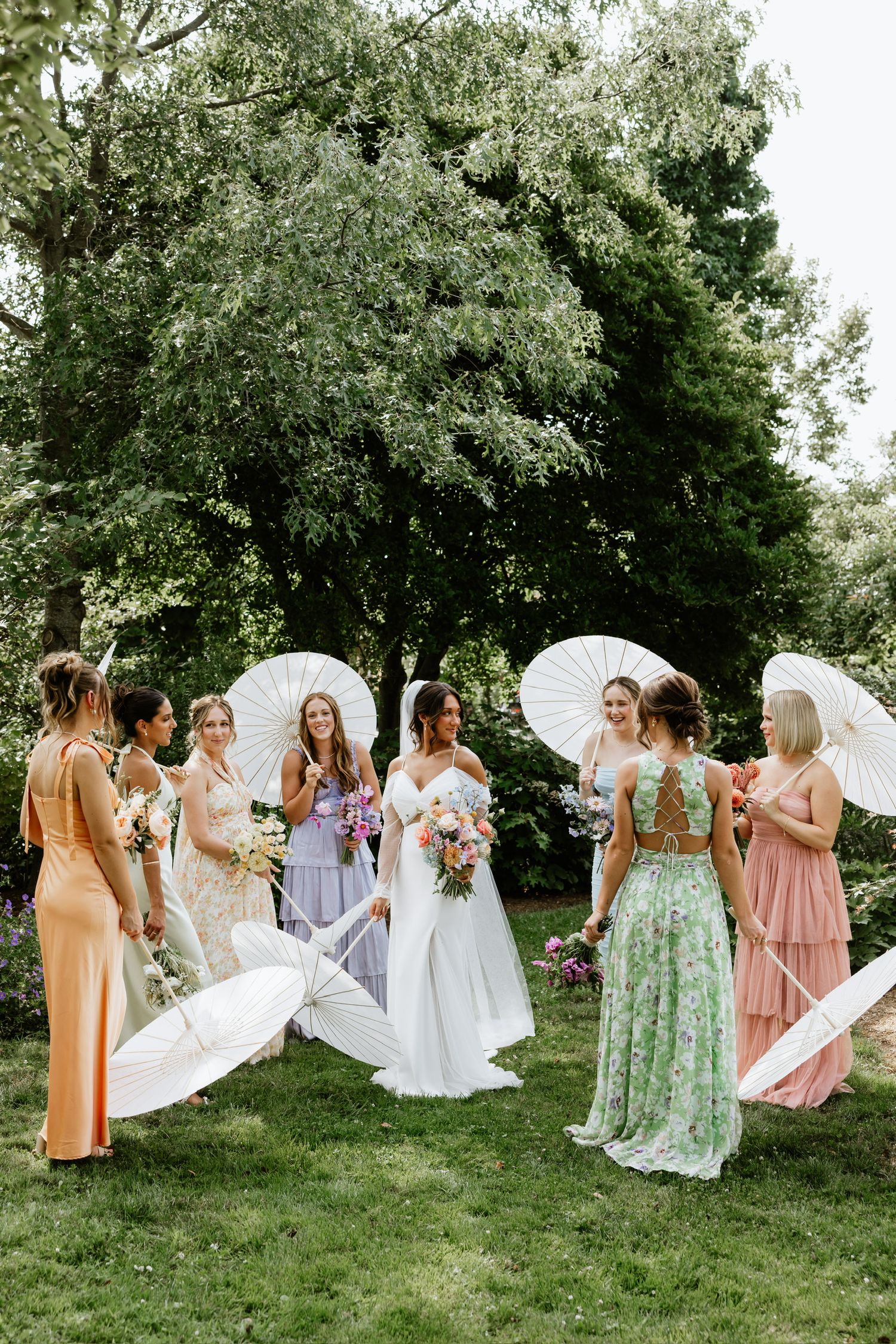 People in elegant dresses gathered outdoors for a wedding celebration under mature trees.