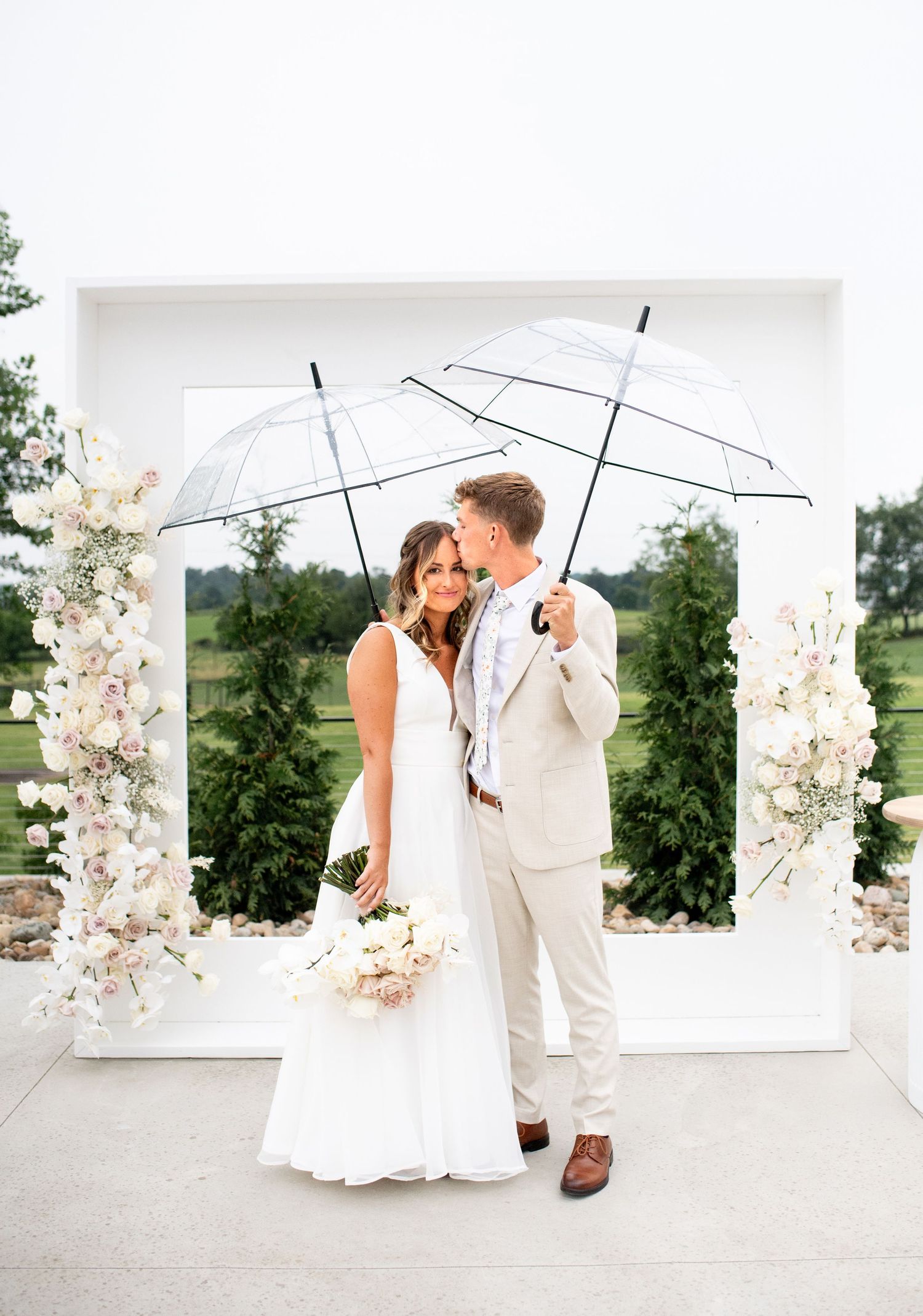 Wedding couple poses under clear umbrellas by a white floral arch in an elegant outdoor setting.