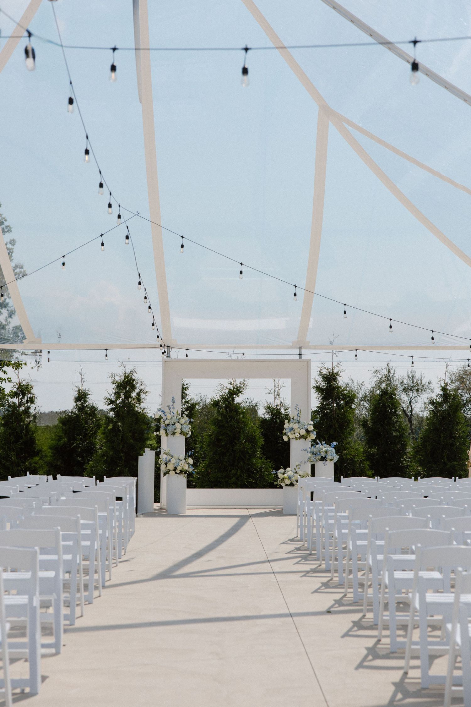 Clear tent wedding ceremony setup with white chairs and string lights creating aisle to floral altar.