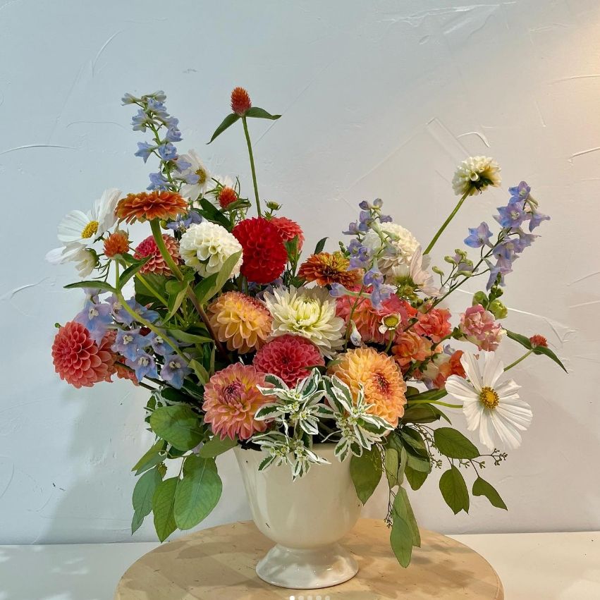 Vibrant summer arrangement with orange dahlias, white daisies, and blue delphinium in a cream pedestal vase.