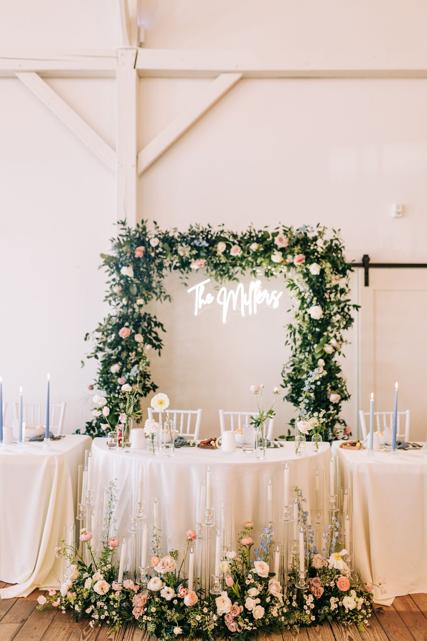 Elegant white wedding sweetheart table with floral arch and neon sign under white beamed ceiling.