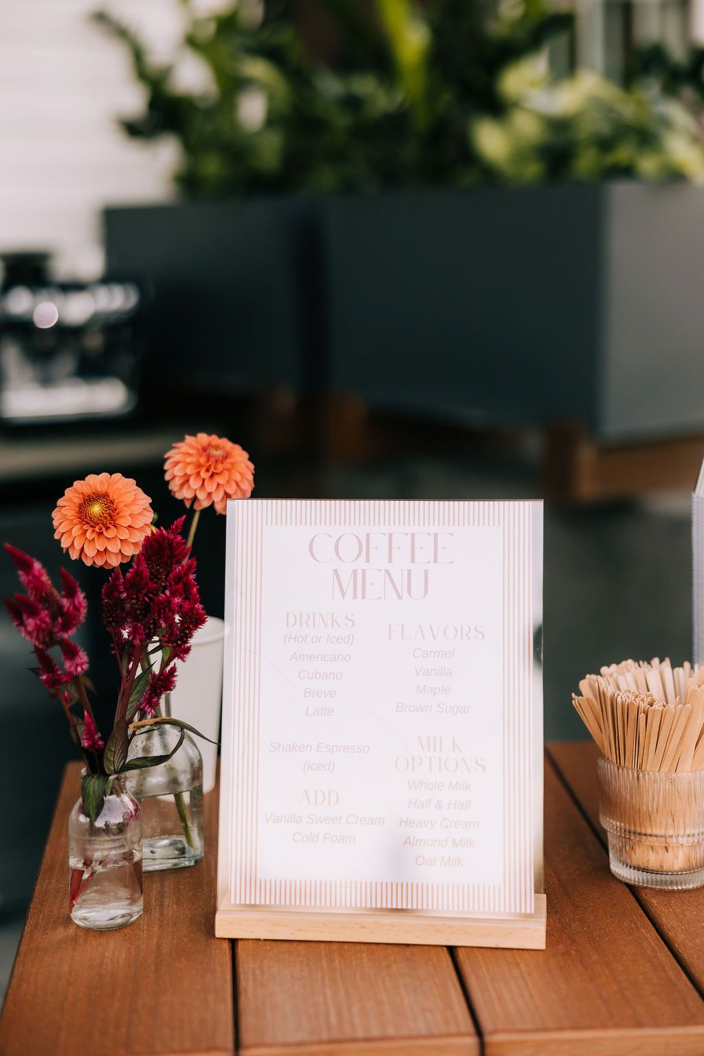 Small vases with pink and burgundy flowers sit beside white place cards on a wooden table at an event.