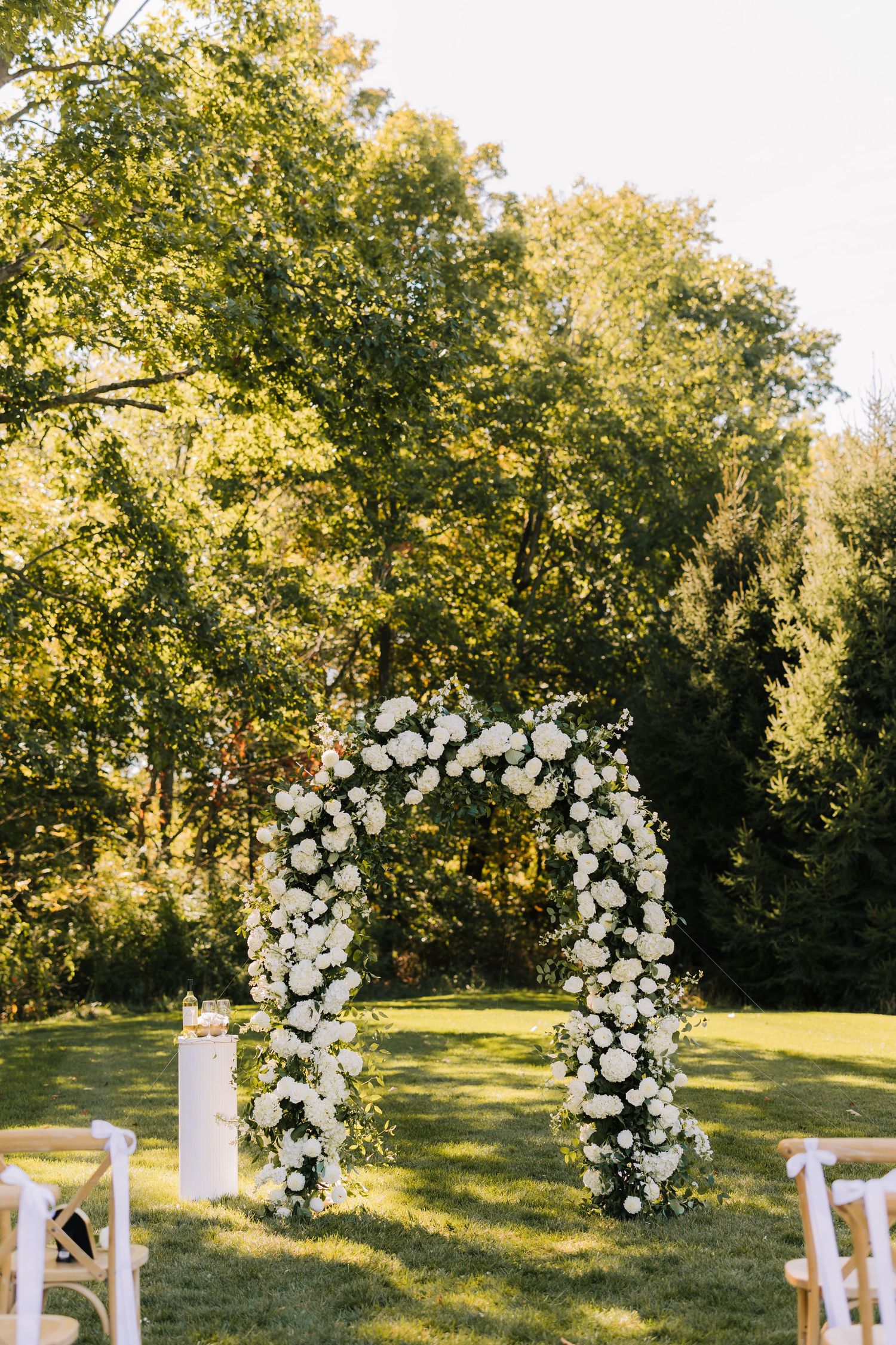A white floral wedding arch decorated with roses and greenery stands on a sunlit grassy lawn during golden hour.