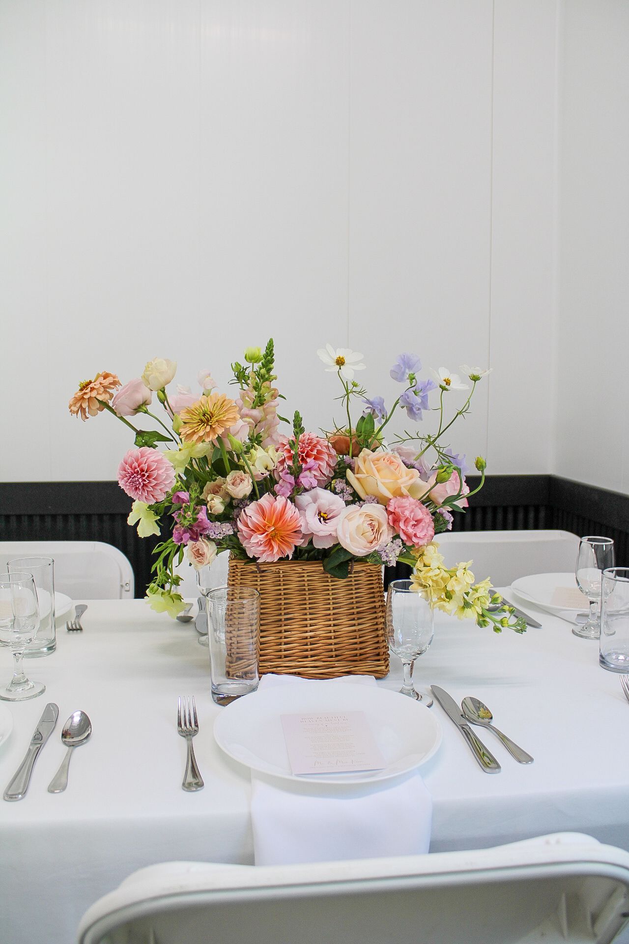 A wicker basket centerpiece filled with colorful spring flowers sits on an elegant white tablecloth with formal place settings.