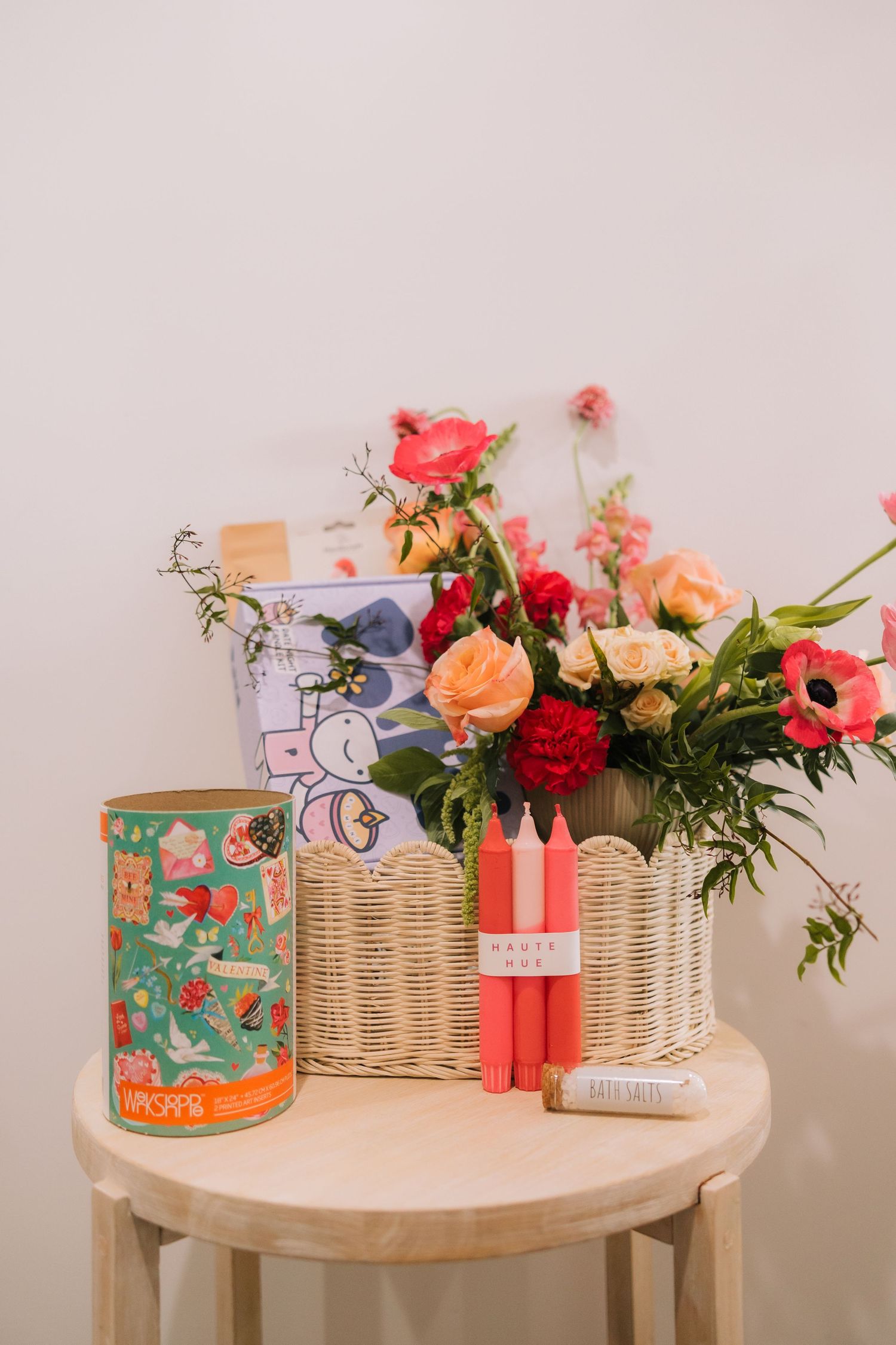 A wooden side table displays a colorful floral arrangement in a wicker basket, alongside red candles and a decorative tin.