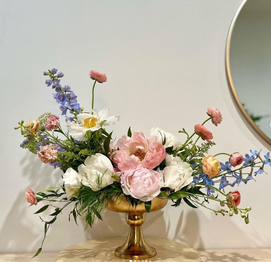 Classic floral design with pink peonies, white roses, and blue delphiniums in a gold compote vessel.
