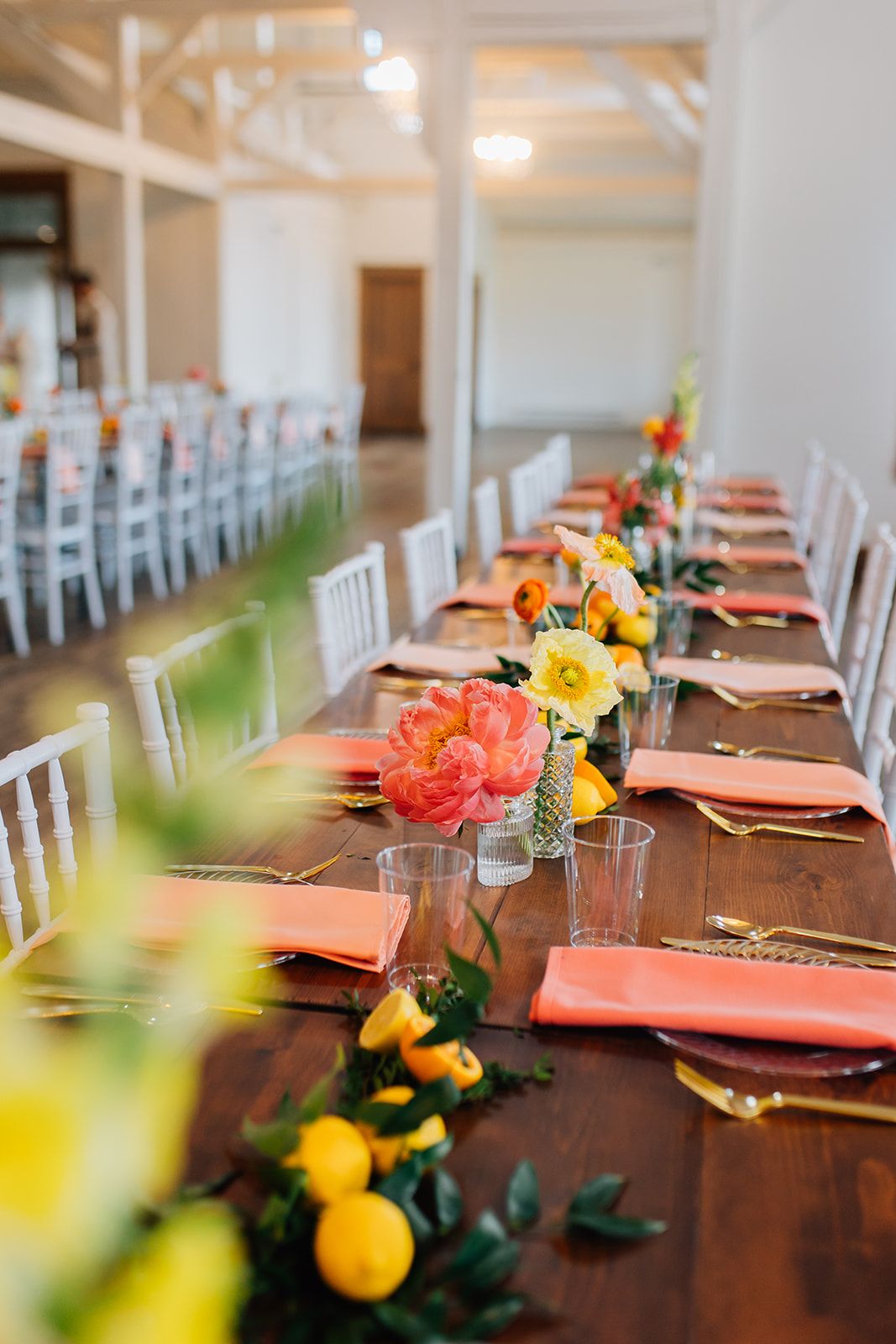 Long wooden dining table decorated with coral napkins, citrus fruits, and colorful floral centerpieces for an elegant event.