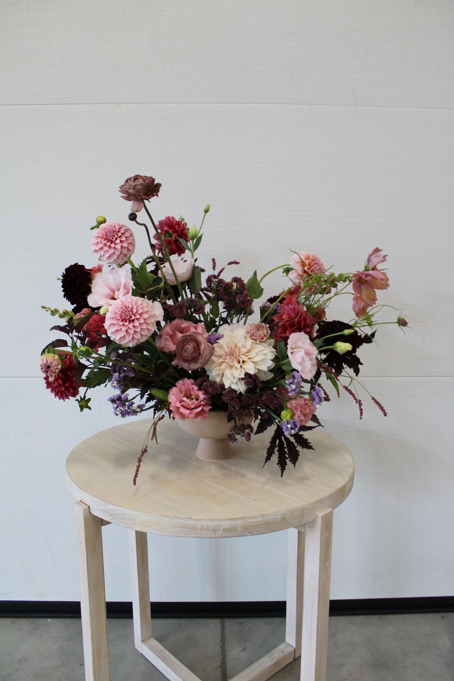 Elegant floral arrangement with pink and burgundy dahlias displayed on a round wooden table against a light gray wall.