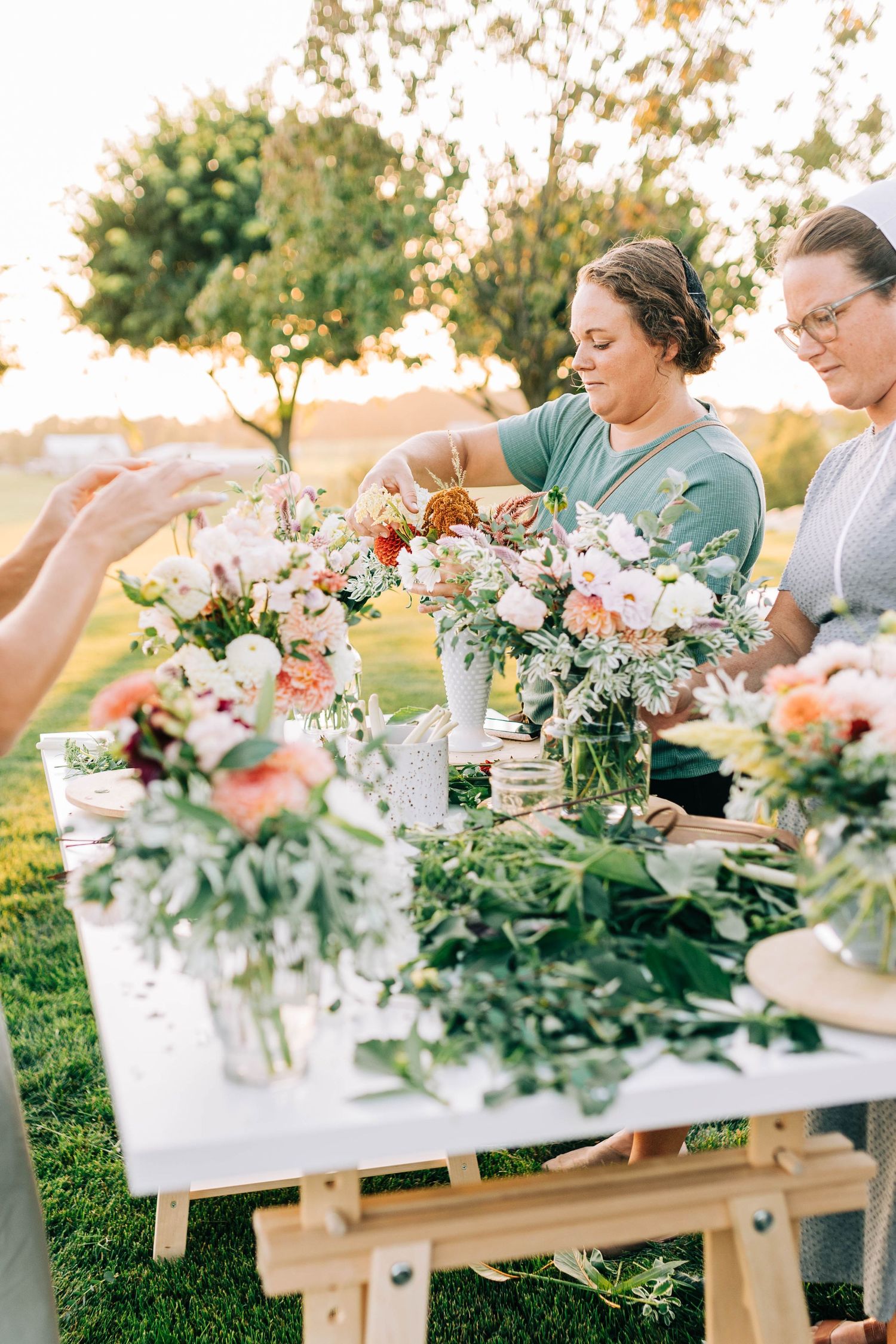 Hands arranging fresh flowers in glass vases on wooden tables during an outdoor workshop.