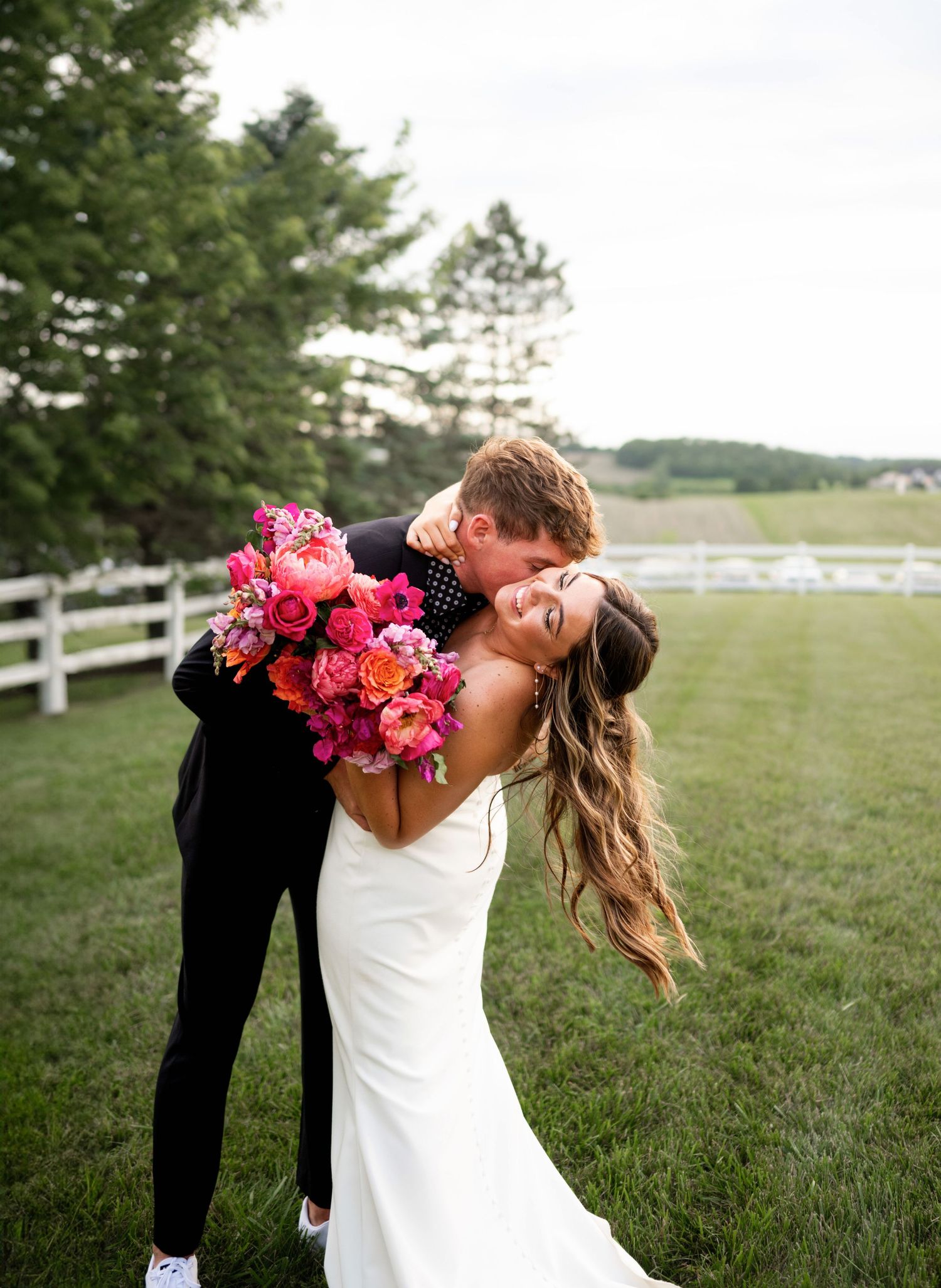Romantic wedding couple embraces in a field with white fence backdrop as bride holds vibrant pink bouquet.
