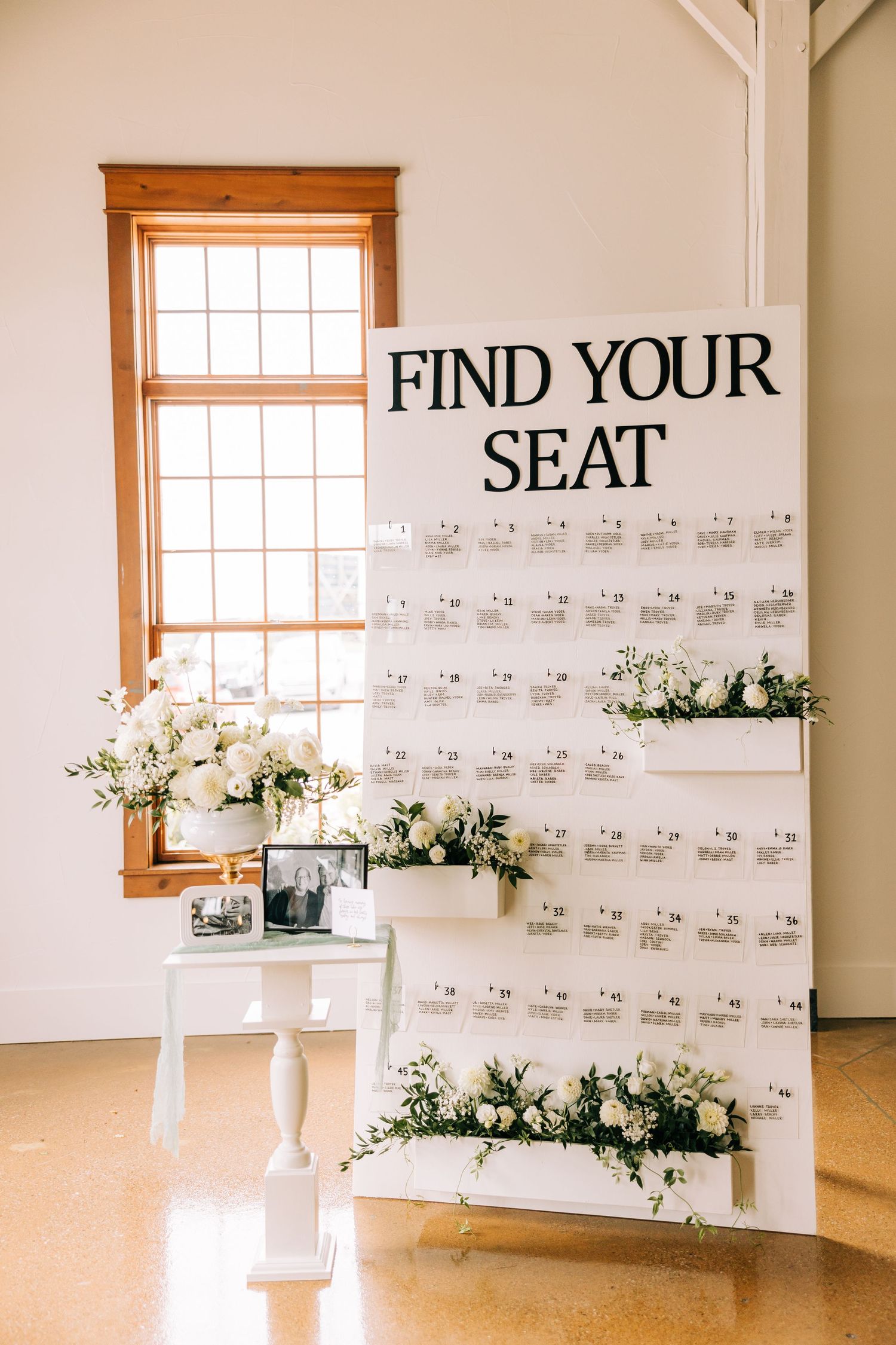 Wedding seating chart display with 'Find Your Seat' text and floral accents next to a sunlit window.