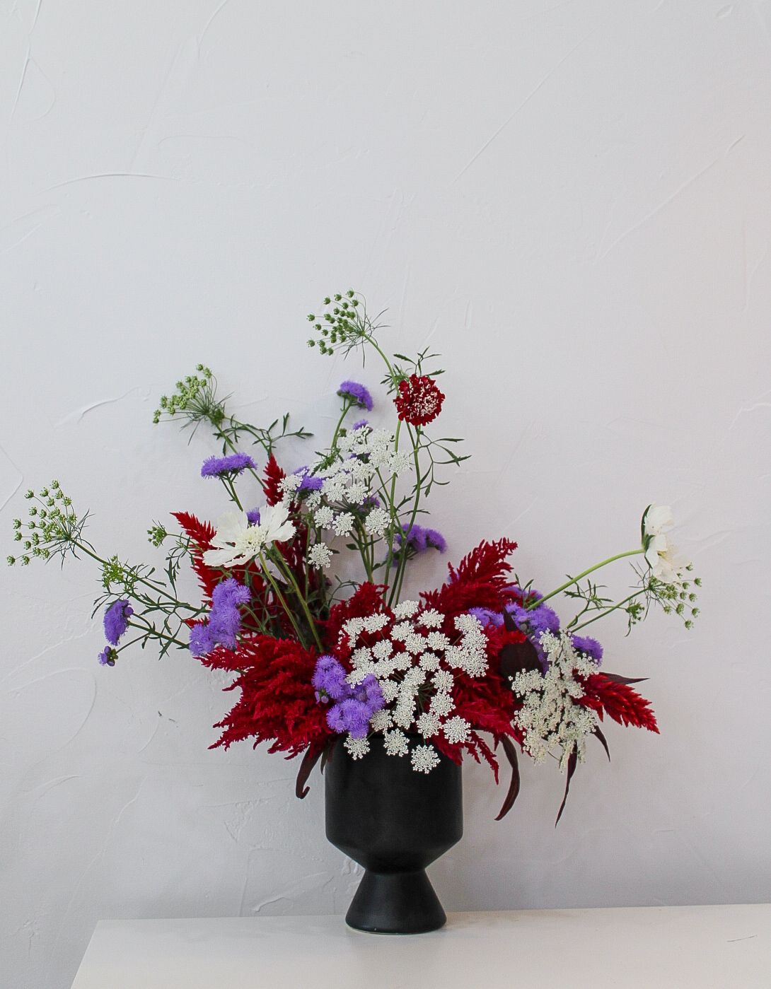 Dramatic floral design with purple blooms, red foliage and white baby's breath in black pedestal vase.