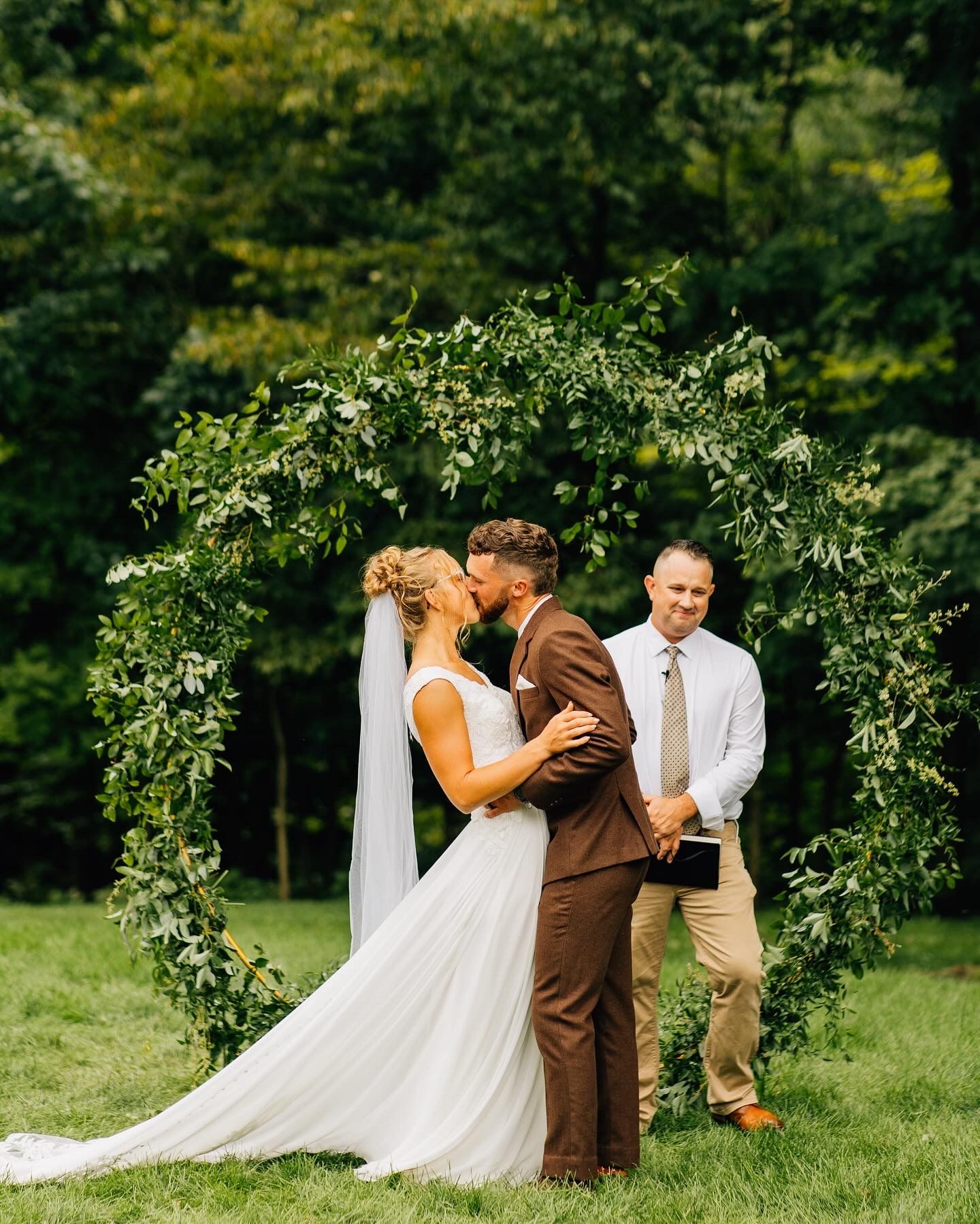 Outdoor wedding ceremony beneath a lush greenery arch in a garden setting.
