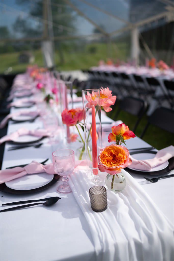 Long reception table with pink and coral flower centerpieces and place settings under clear tent.