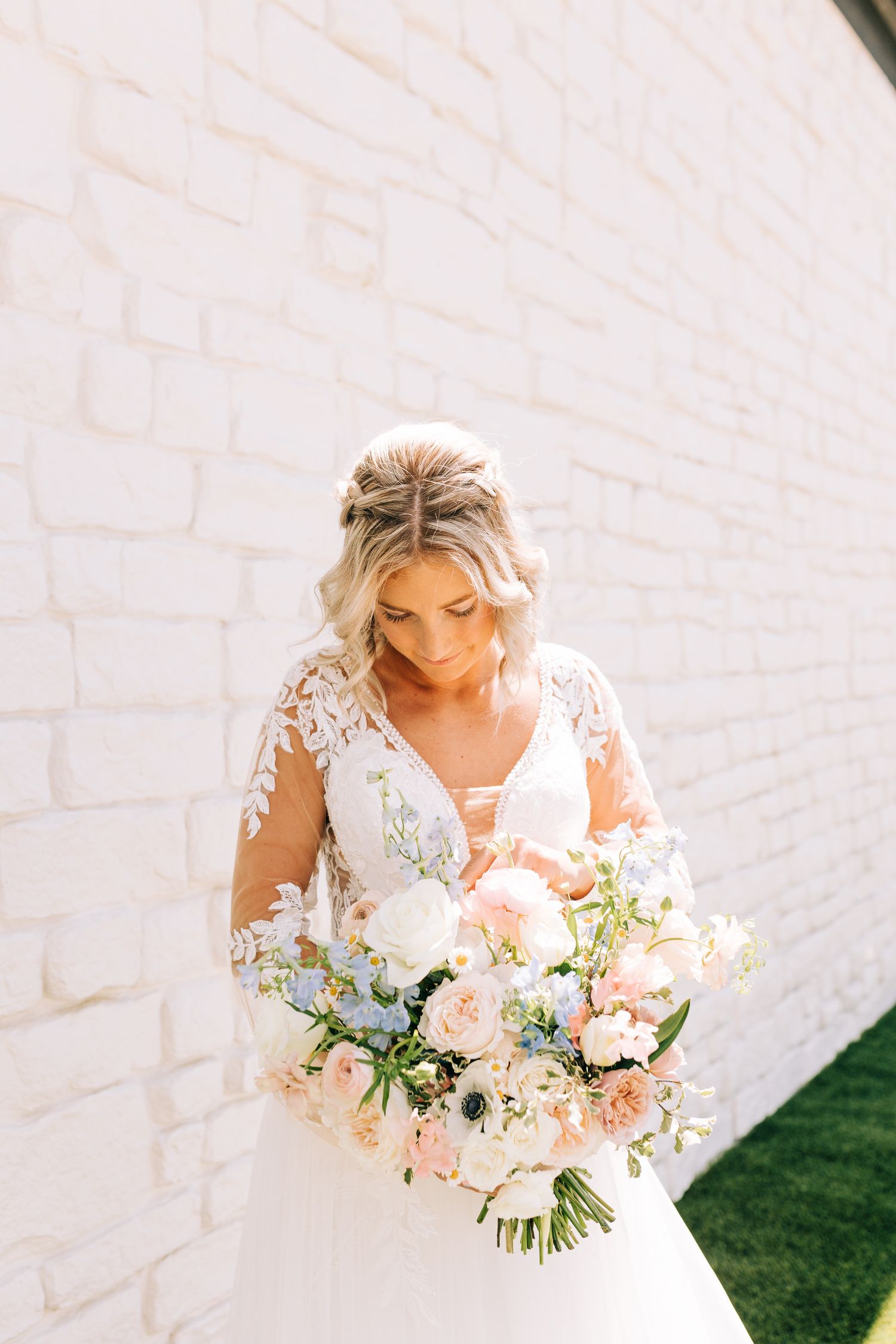 A bride in a lace wedding gown holds a lush bouquet of pink and white flowers against a white brick wall.