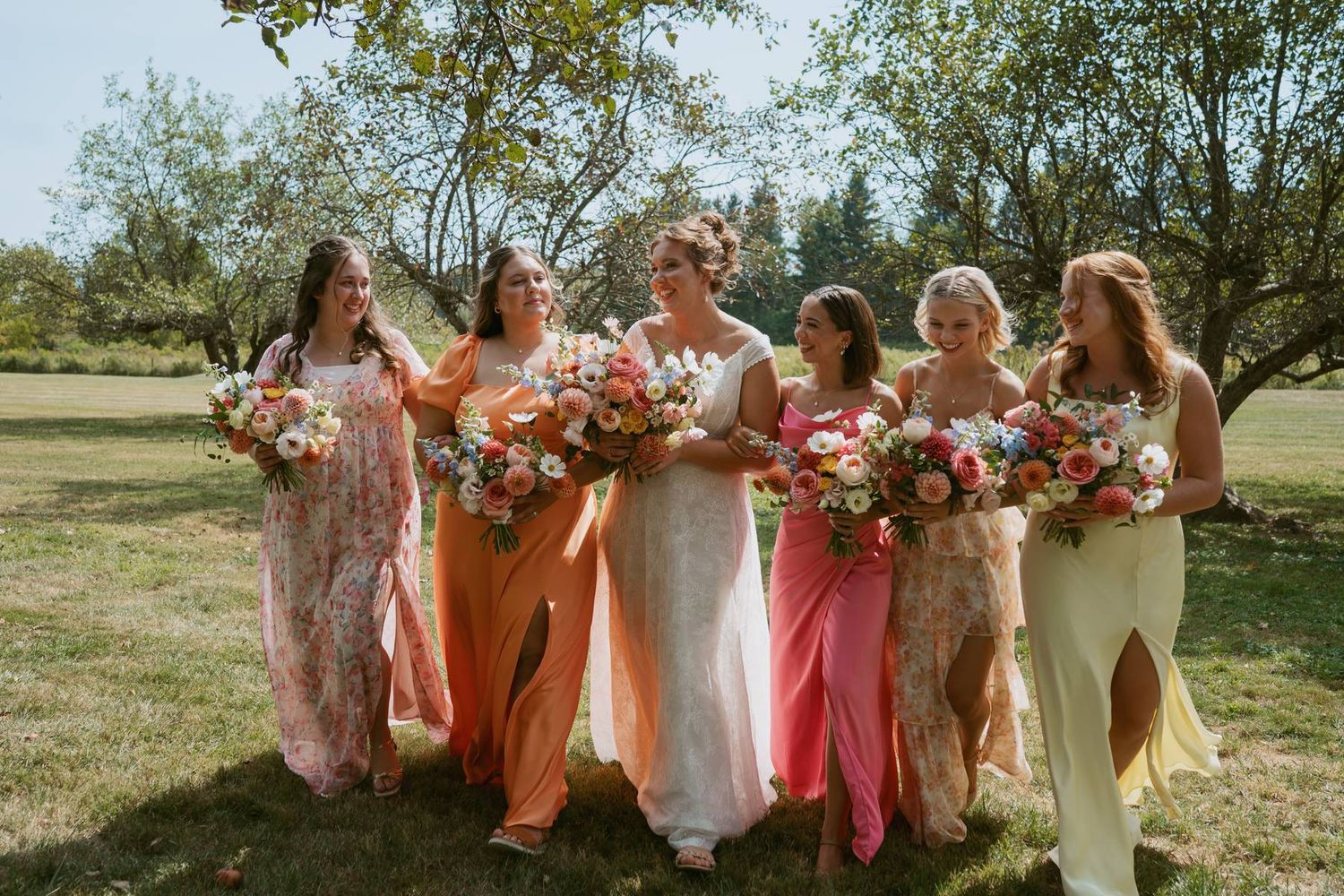A group of bridesmaids in peach and coral dresses pose together in a sunny outdoor setting.