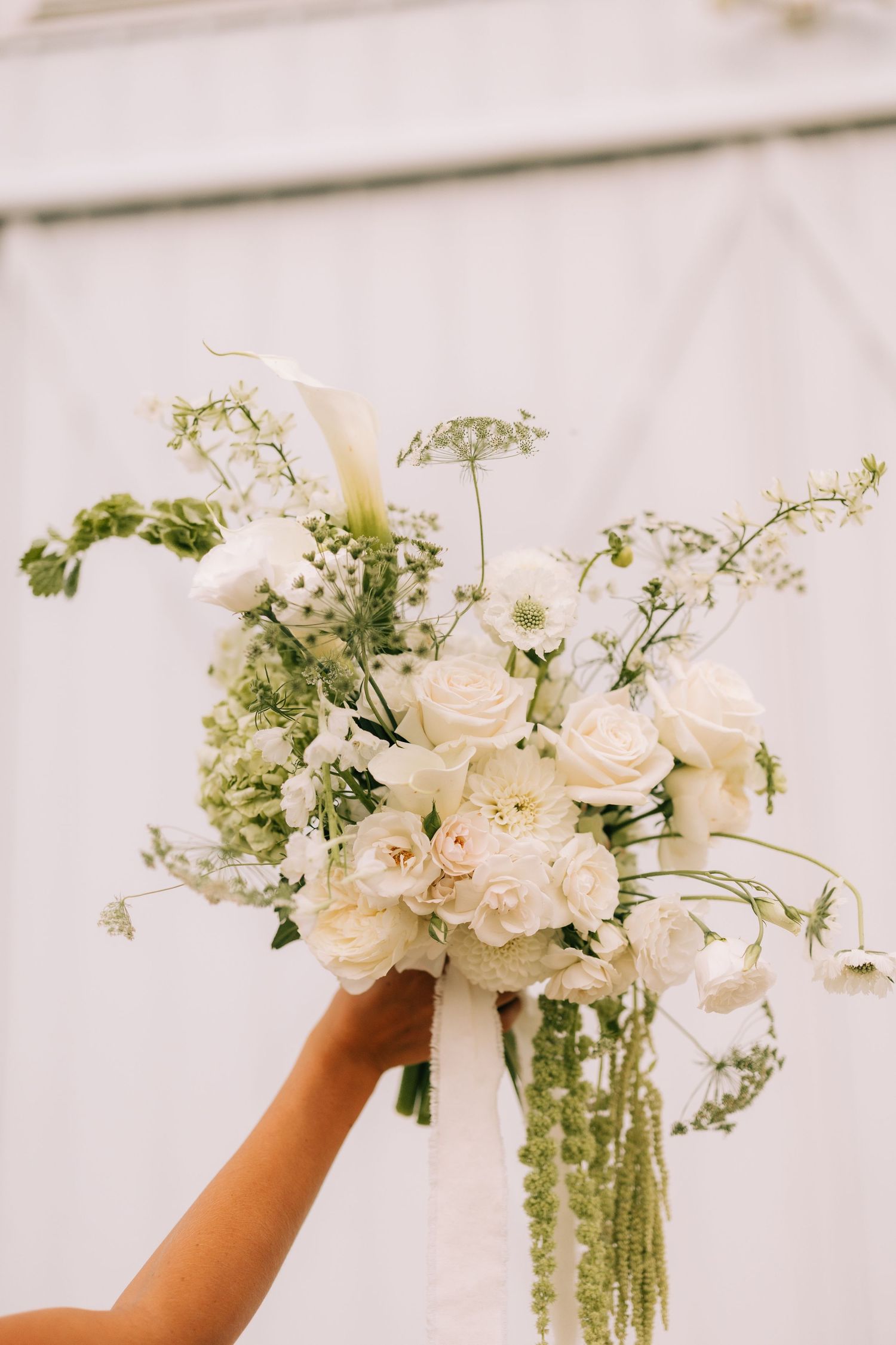 Hand reaching up to arrange delicate white roses and greenery in a tall glass vase against a light background.