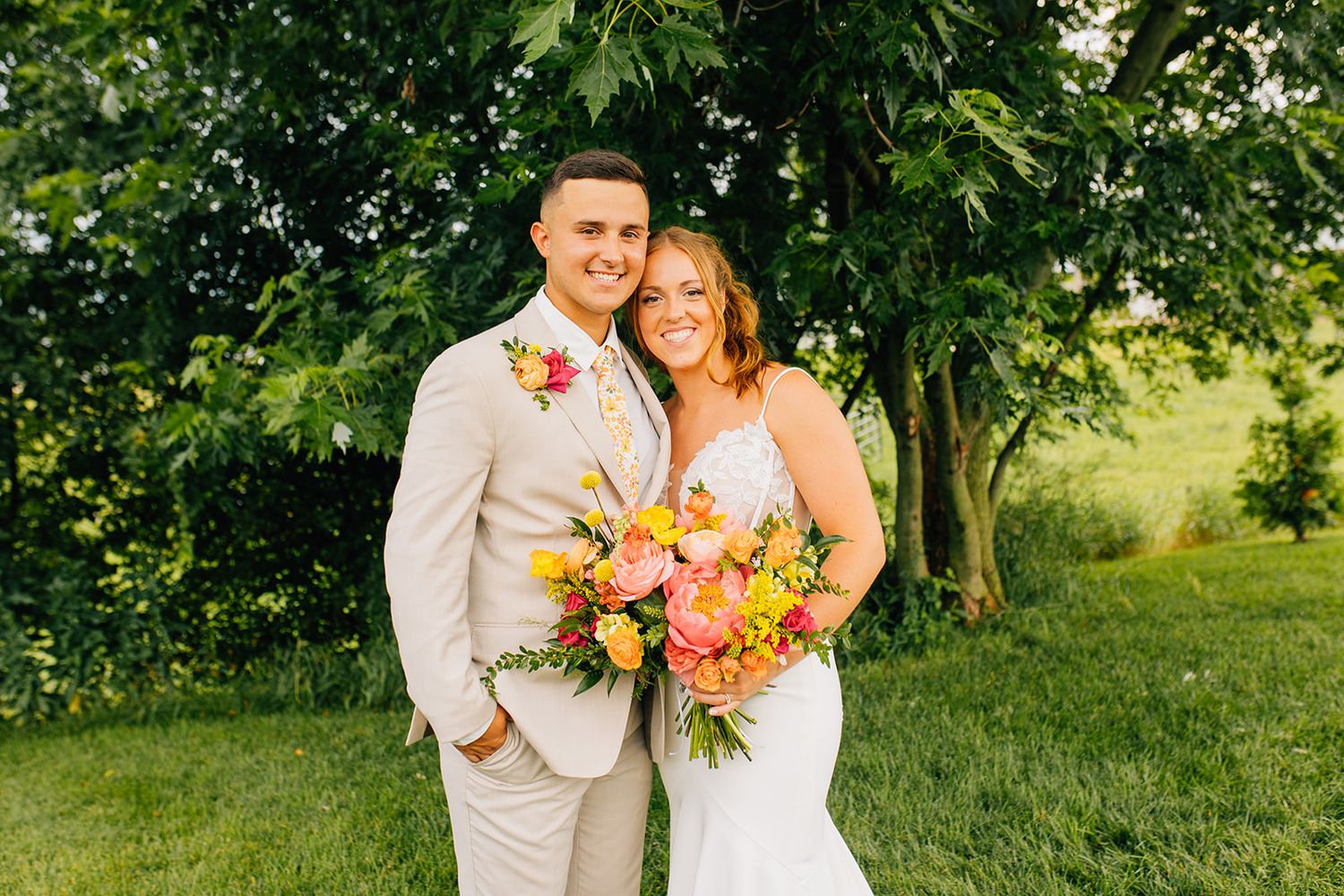 A joyful wedding couple poses outdoors in a lush garden setting with vibrant summer greenery and colorful bouquet.