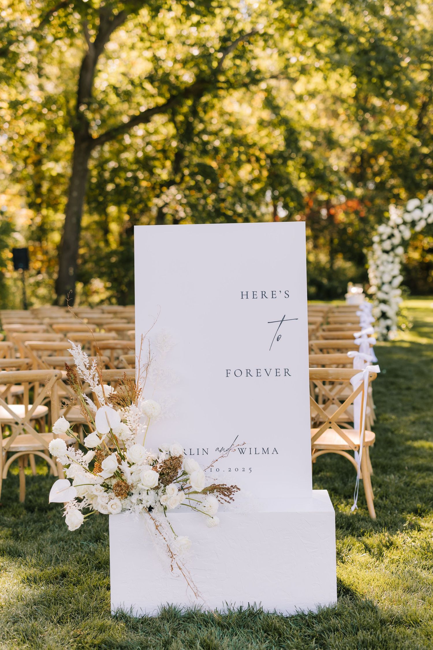 White wedding signs with floral decorations displayed on wooden chairs in a sunny outdoor garden setting.