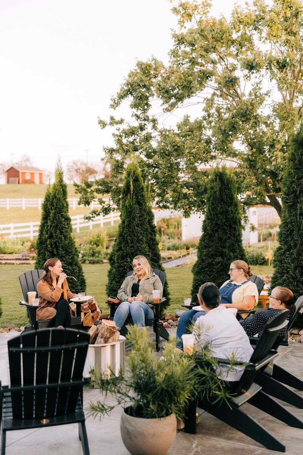A group of people relax in black Adirondack chairs on a patio overlooking a scenic landscape at sunset.