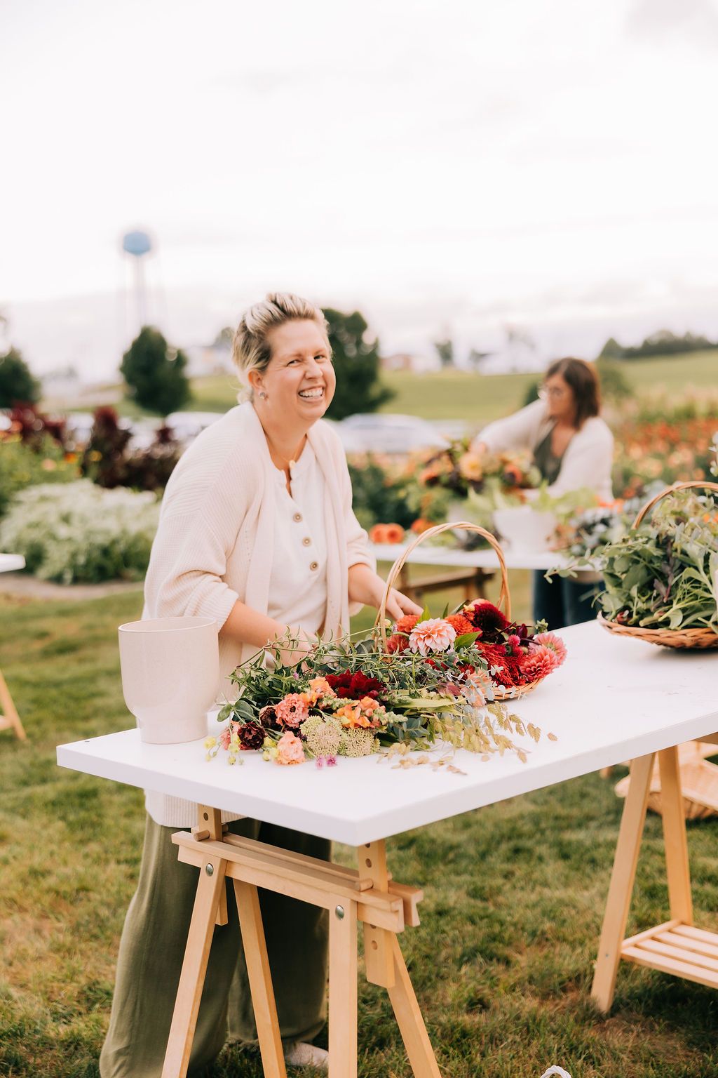 A florist arranges flowers and greenery on a white table at an outdoor event venue.