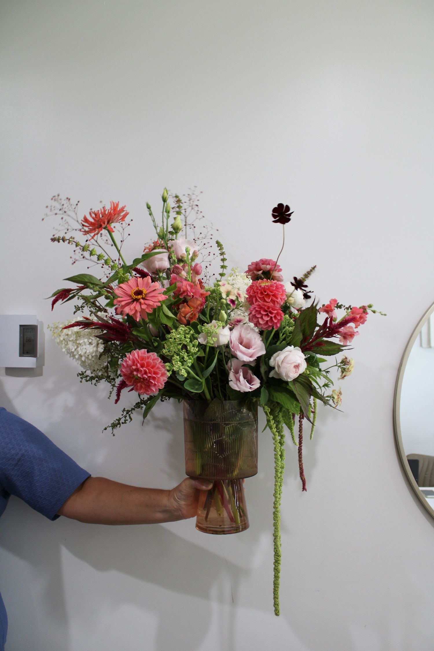 A hand holds a vintage glass vase filled with pink and coral flowers including roses, carnations and delicate greenery.