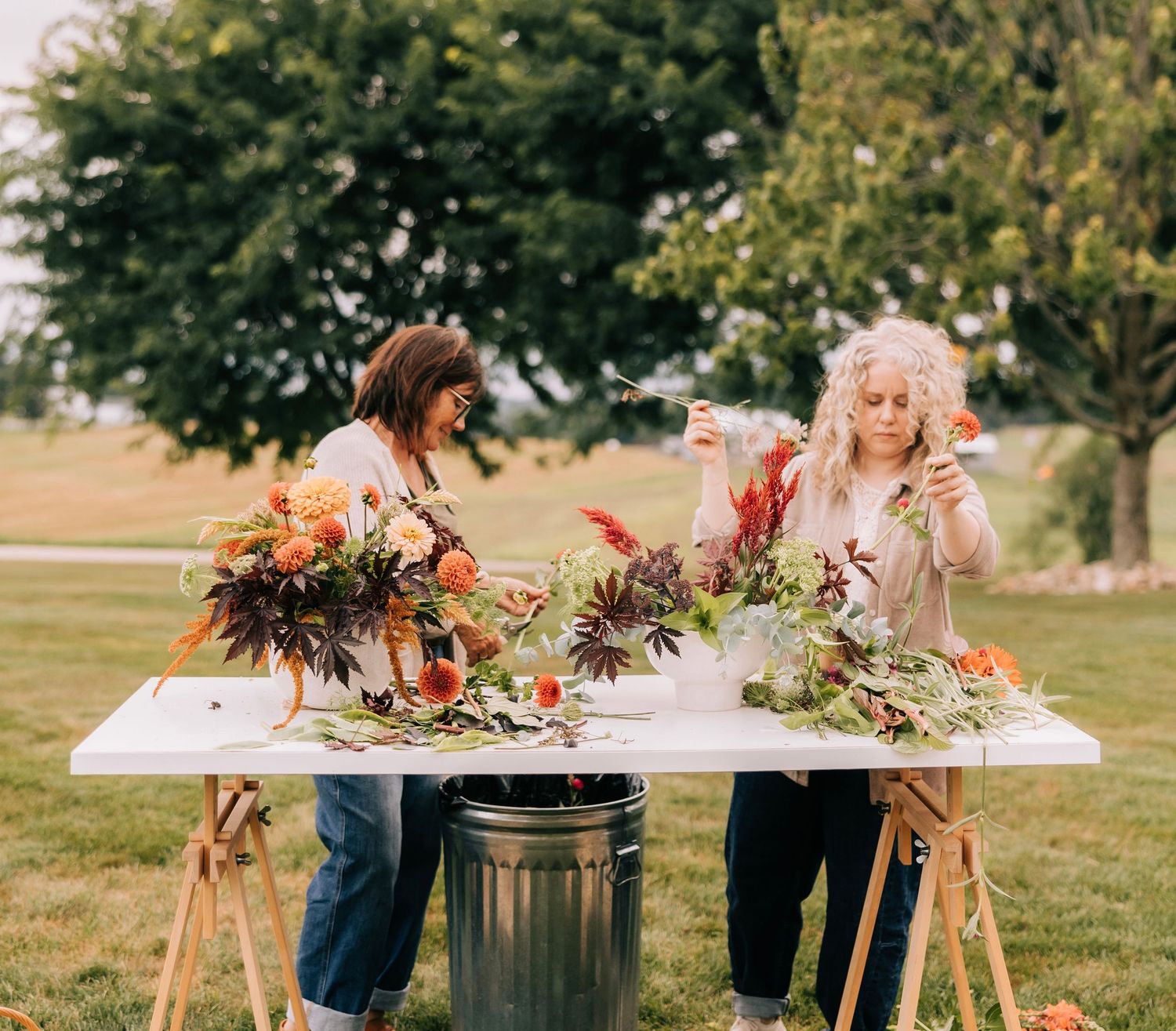 Two women arrange colorful autumn floral bouquets at an outdoor white table surrounded by lush green grass and trees.