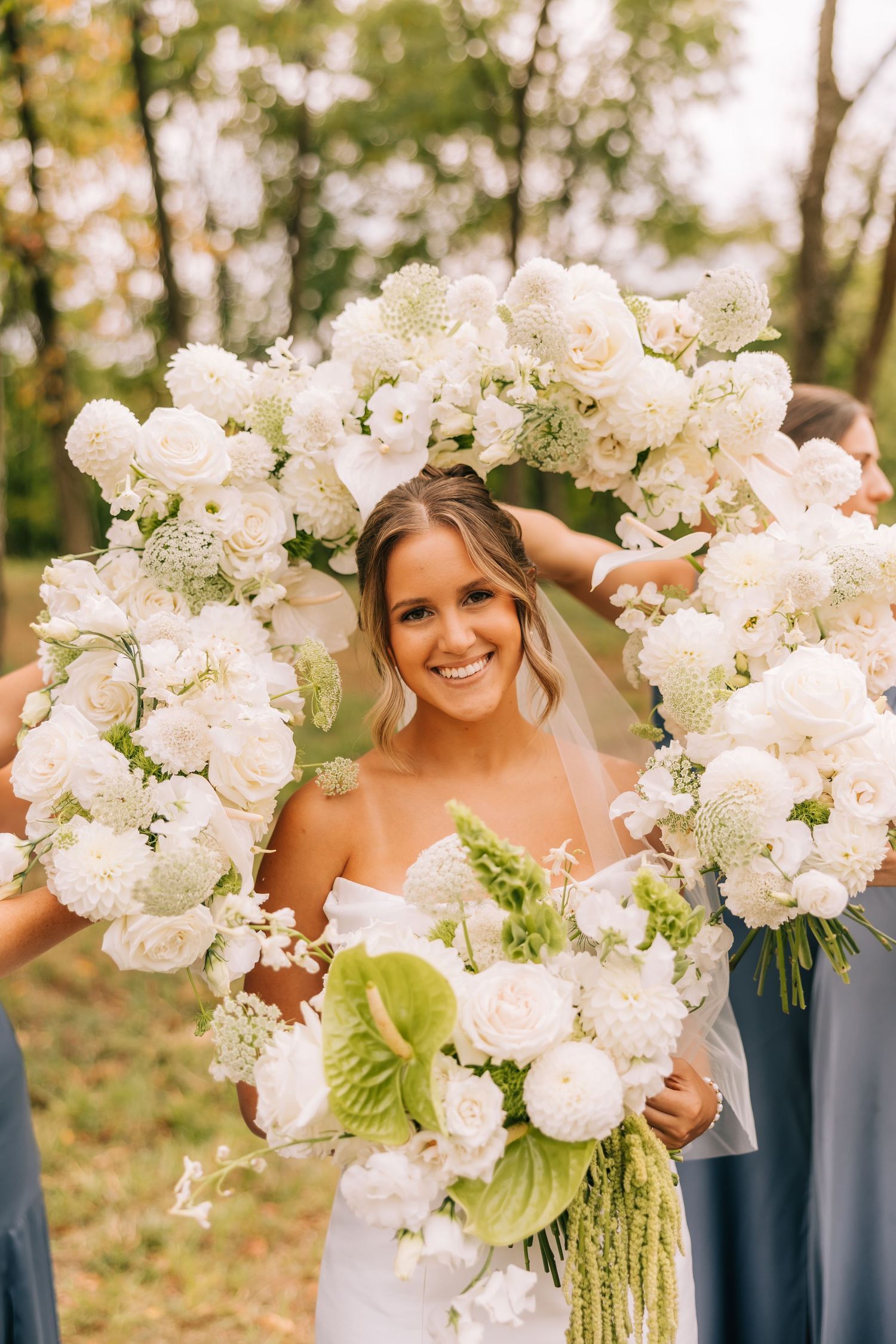 Bridesmaids in blue dresses hold lush white floral bouquets with greenery accents in an outdoor setting.