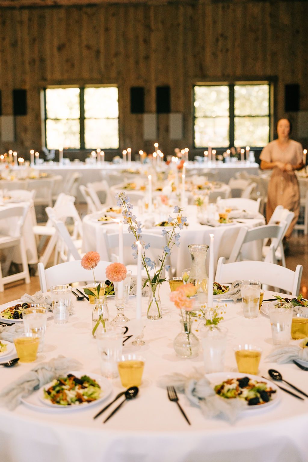 Wedding reception tables set with white linens, glassware, and candles in a rustic barn venue.