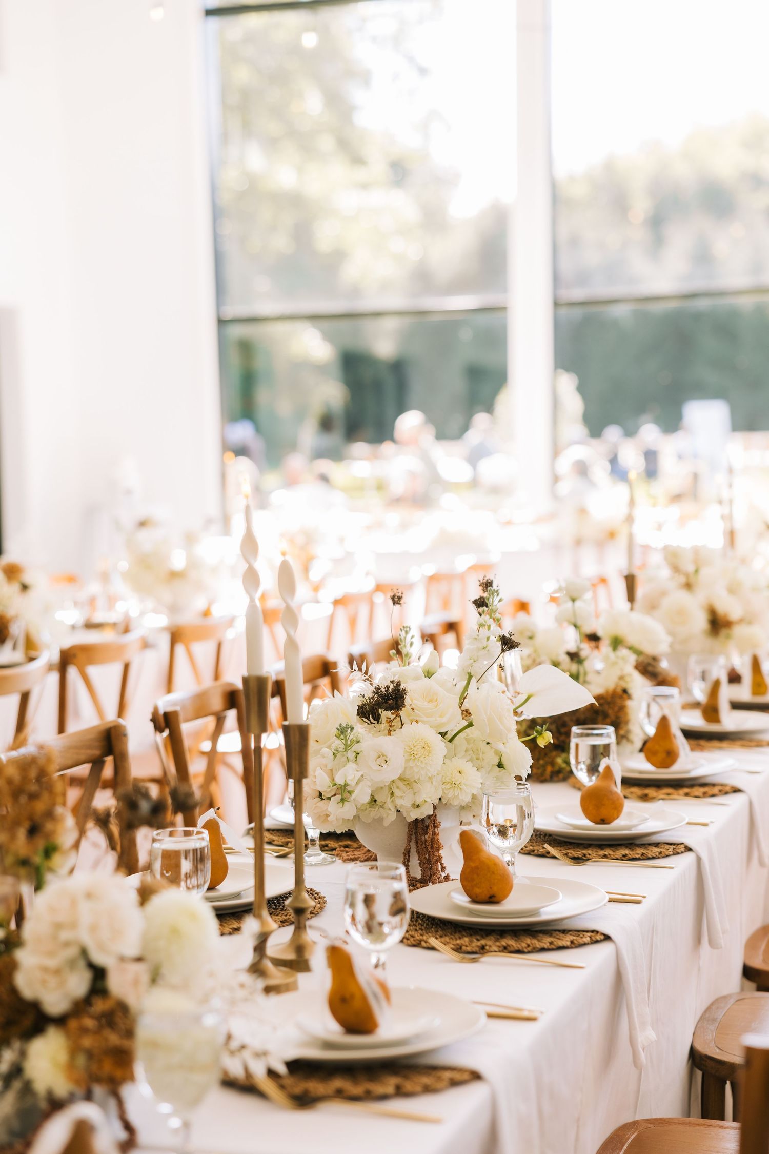 Elegant wedding reception tables set with white linens, gold accents, pears, and white floral centerpieces in a bright venue.