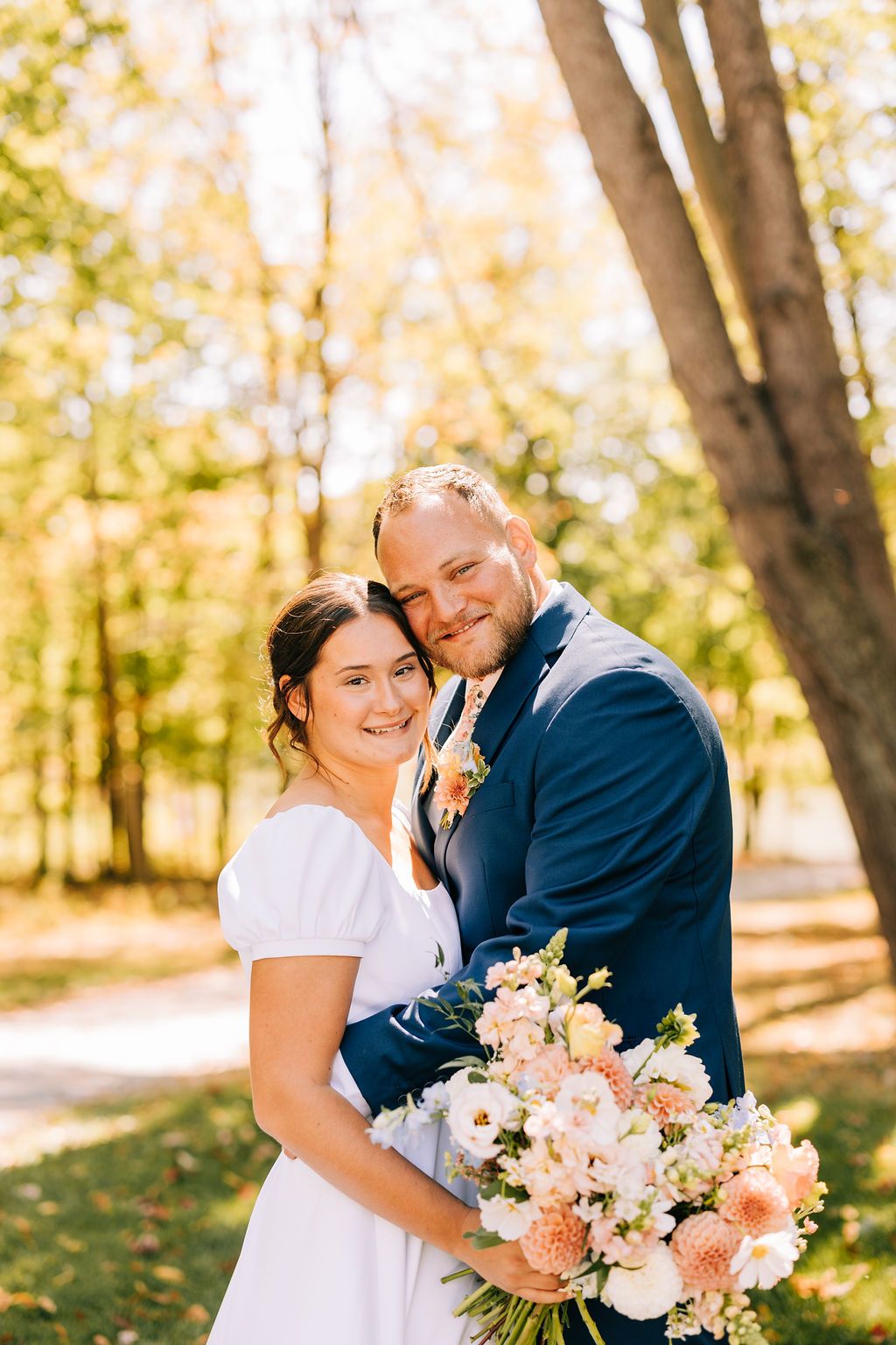 Happy couple embraces in a sun-dappled autumn forest setting while holding a pastel floral bouquet.