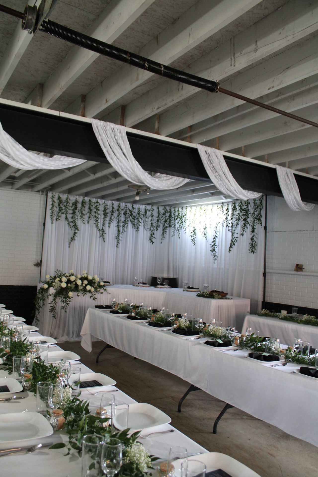 Modern wedding reception space with white draping and hanging greenery above long dining tables.