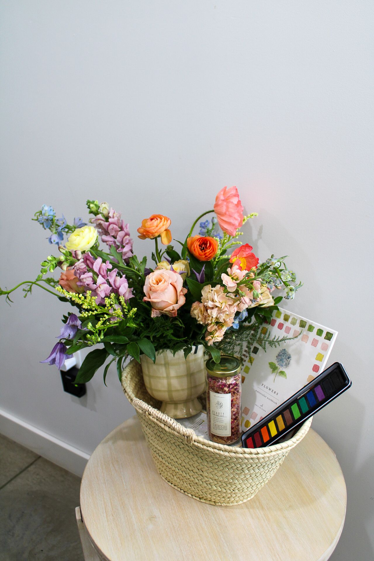 Colorful floral arrangement in a woven basket with watercolor paints and botanical book on a wooden table.