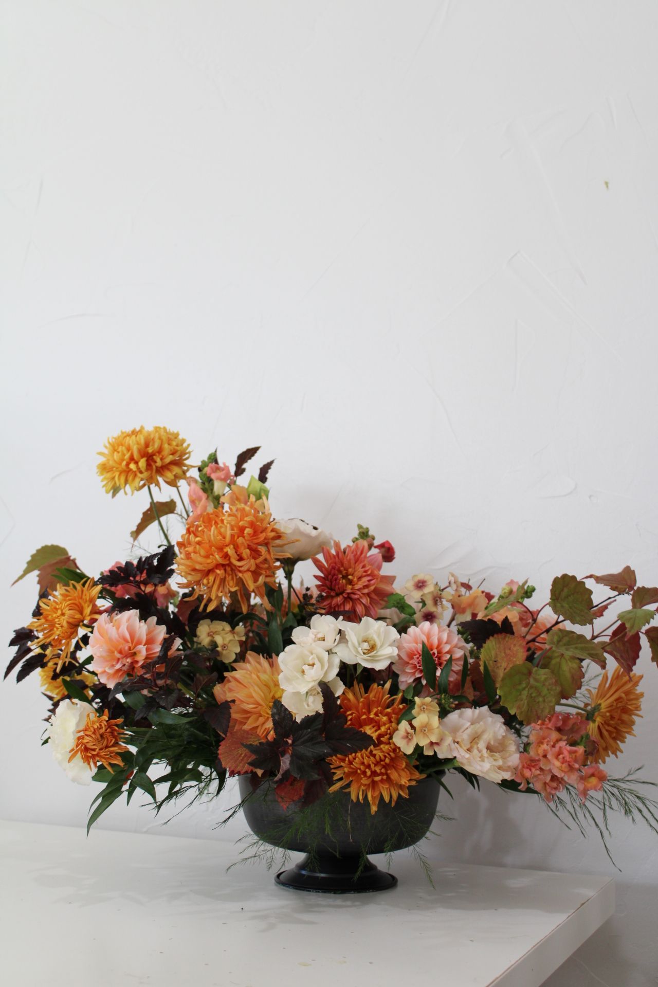 Fall-themed flower arrangement featuring orange dahlias, roses and greenery in a dark pedestal vase.