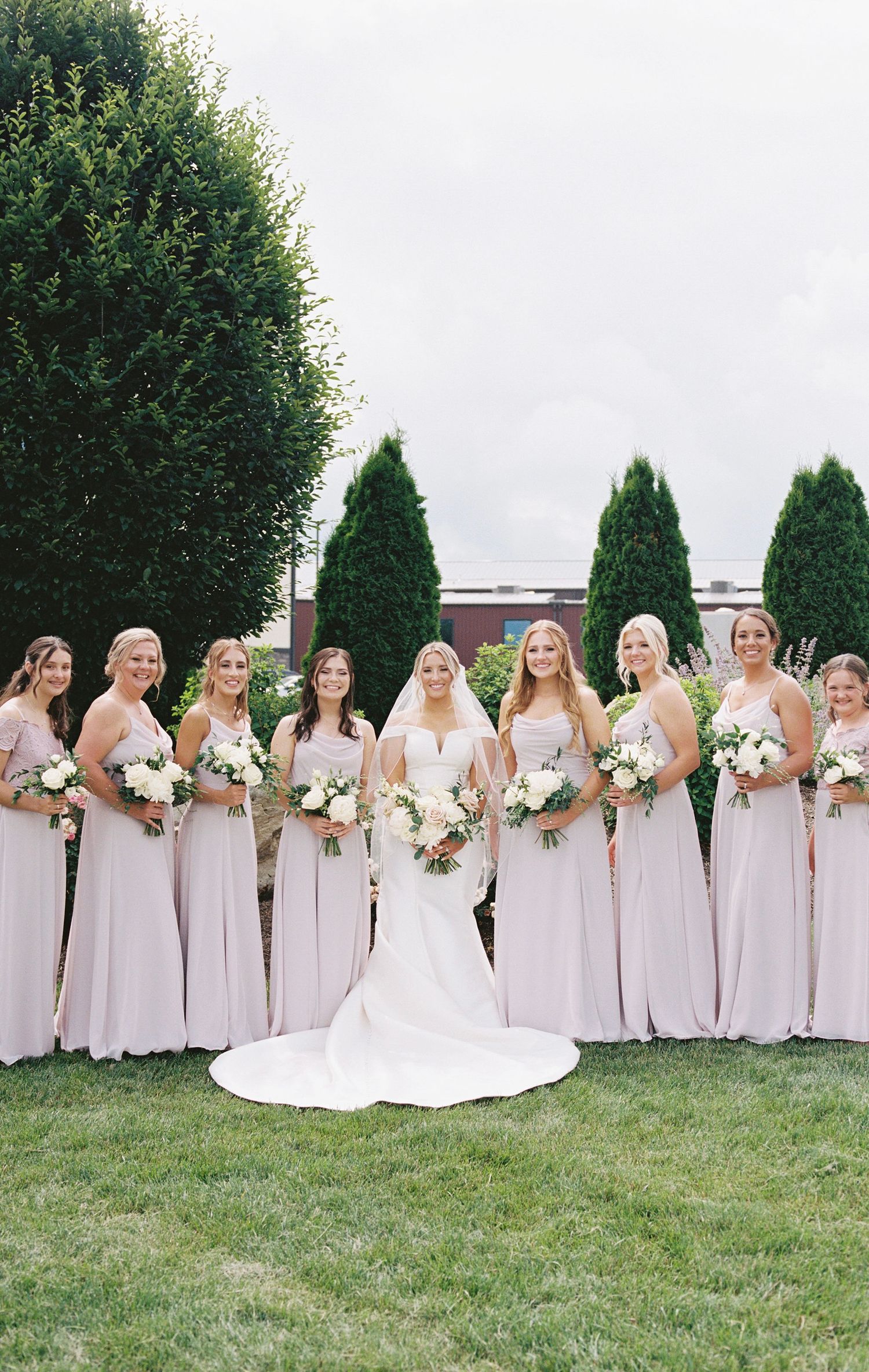 Bridesmaids in pale pink dresses stand with bride against hedge backdrop.