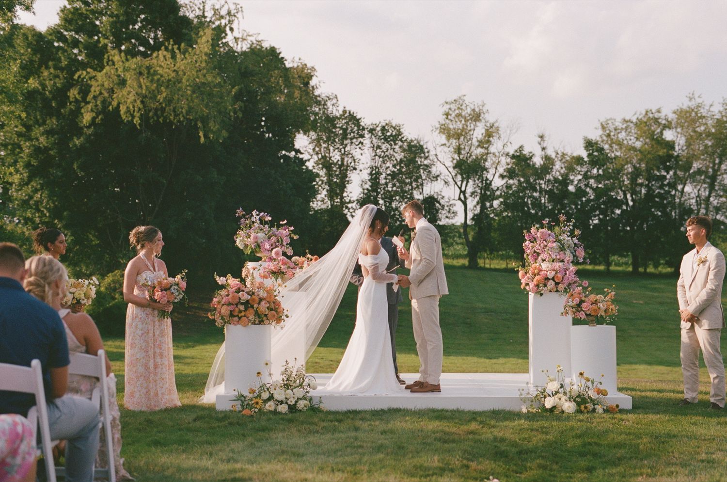 Outdoor wedding ceremony setup with floral pillars and white chairs on green lawn.