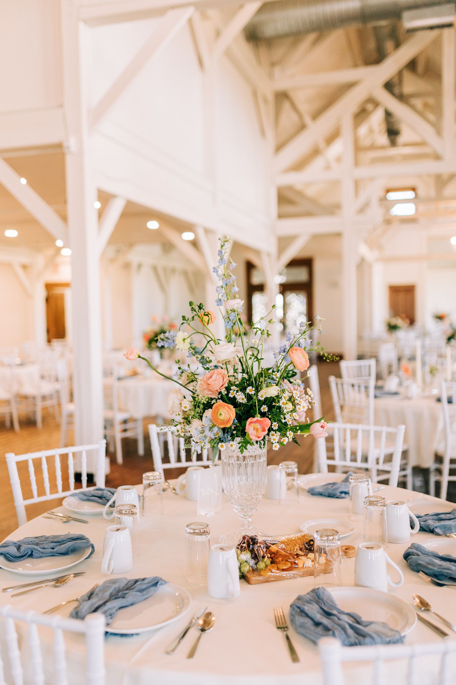 Bright spring wedding table setting with peach floral centerpiece and blue napkins in white venue.