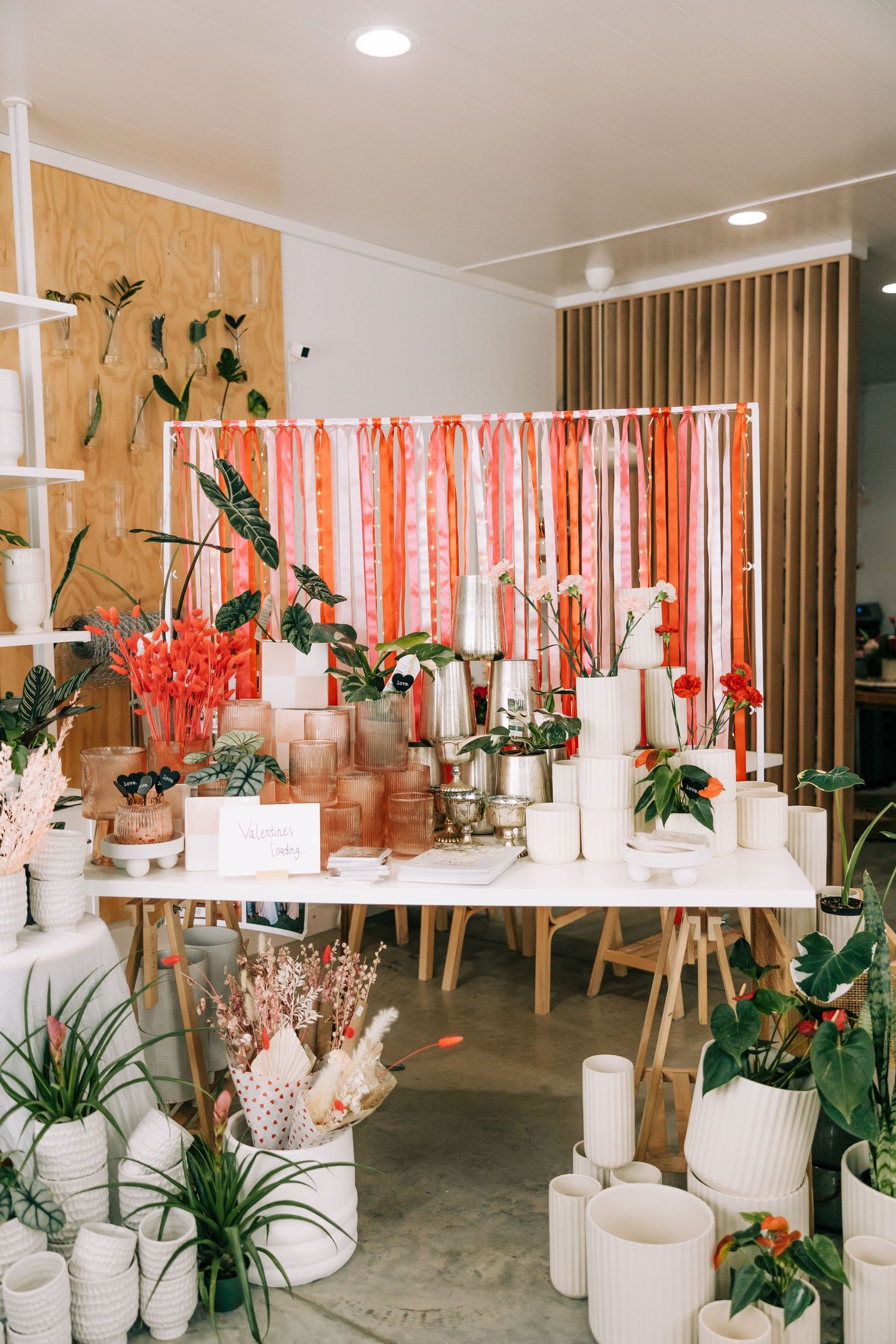 Modern retail display with white ceramics, plants and coral streamers against wood panel wall.