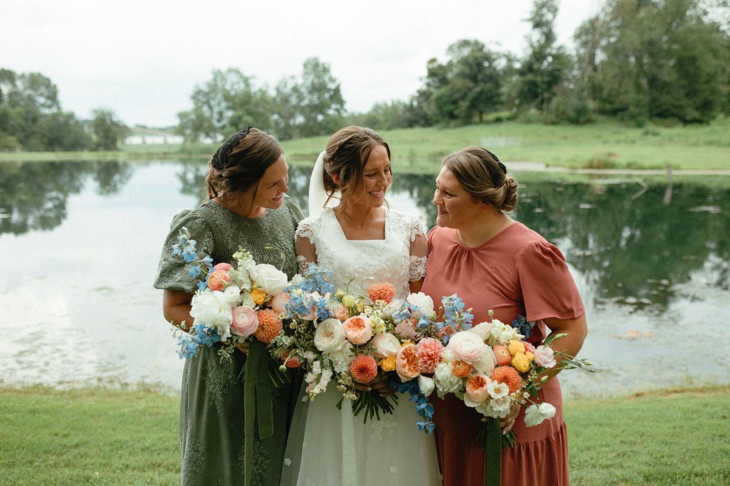 A bridal party wearing burgundy dresses holds colorful bouquets while standing near a serene lake on a sunny day.