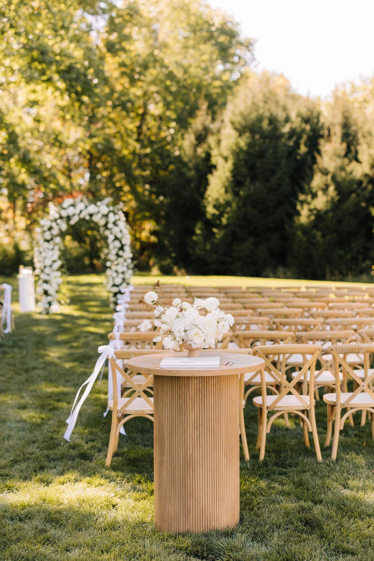 Rustic wooden chairs arranged in rows on a grassy lawn with a circular wooden table decorated with flowers at sunset.