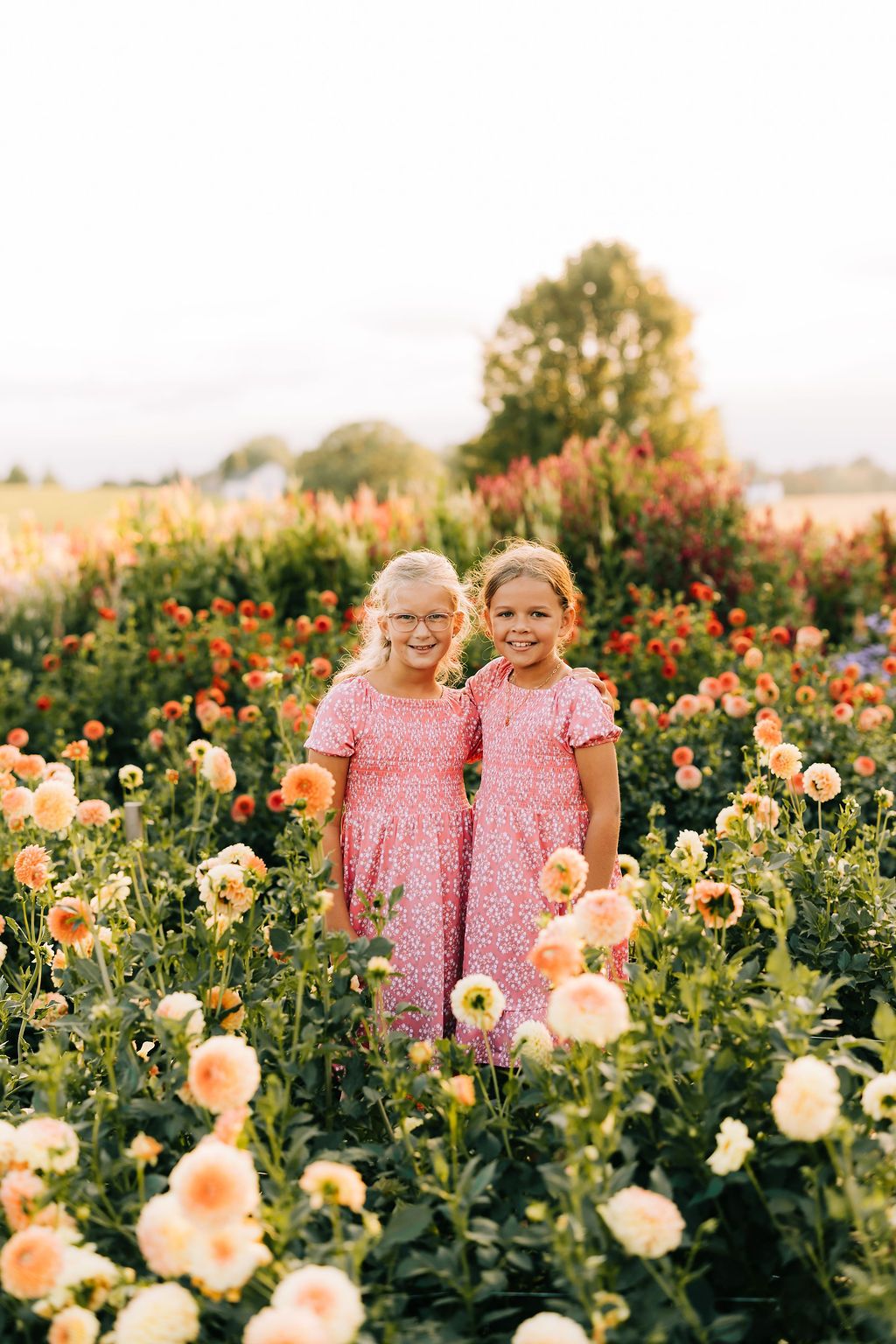 Two people in matching pink floral dresses stand together in a blooming dahlia flower field at sunset.